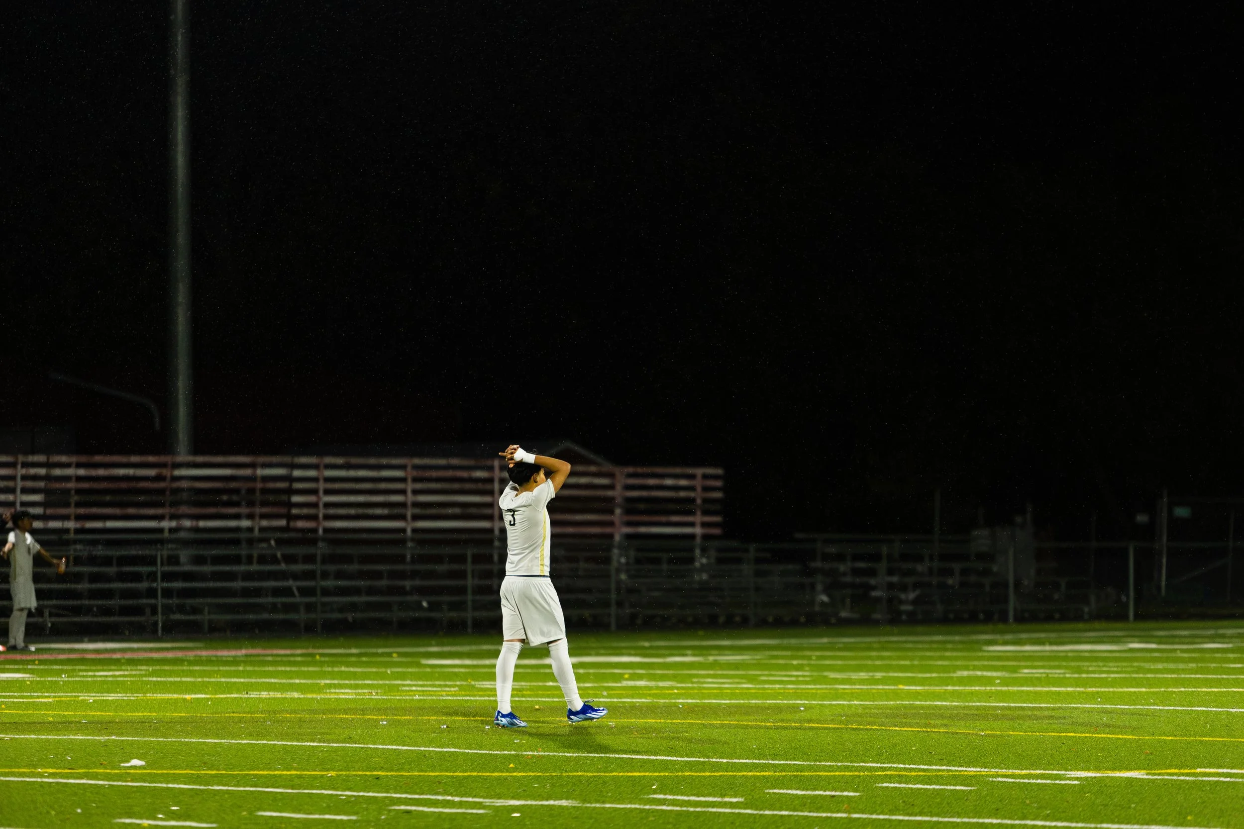 A soccer player stands on a field at night with hands on head, wearing a white uniform and blue cleats. There are empty bleachers and another player in the background.