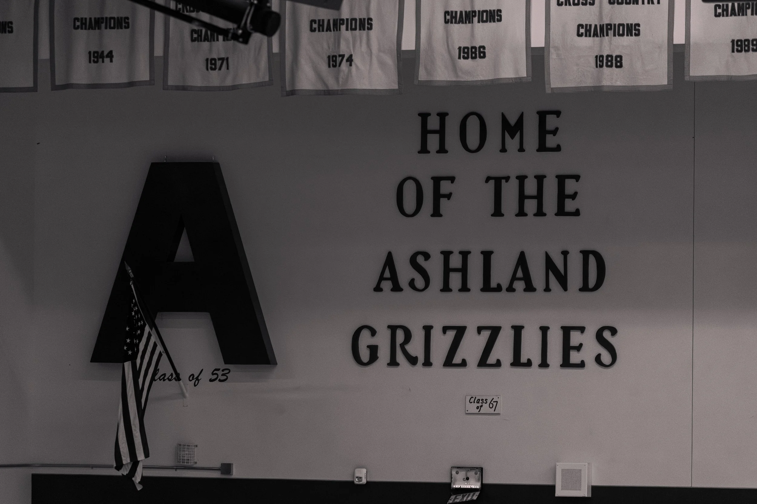 School gym wall with banners celebrating basketball championships and the phrase "Home of the Ashland Grizzlies" in large letters. There is a large black letter A with a small American flag hanging from it.