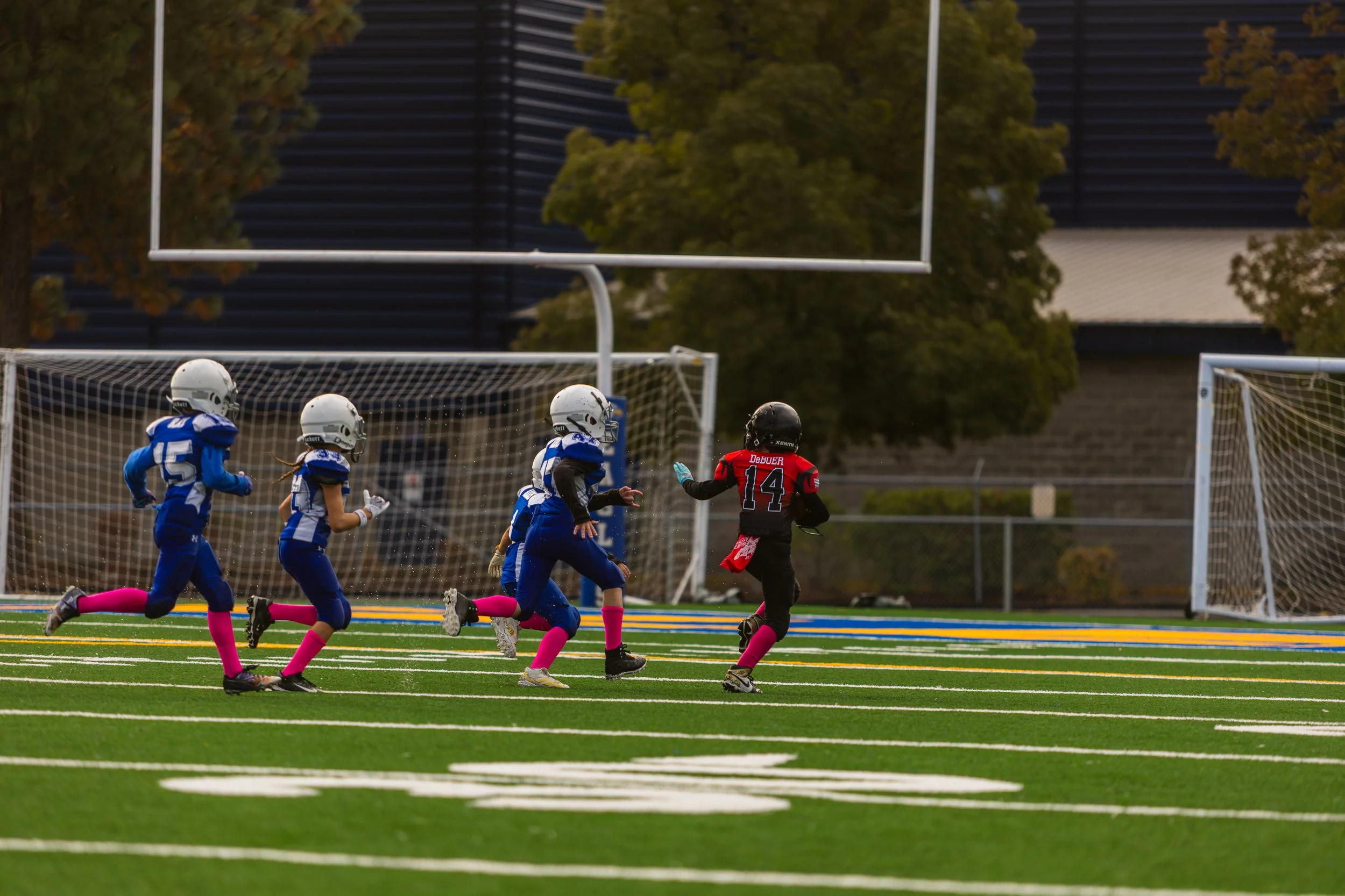 Youth football game on a field, with one child in a red uniform running ahead of children in blue uniforms, all wearing helmets and pink socks.