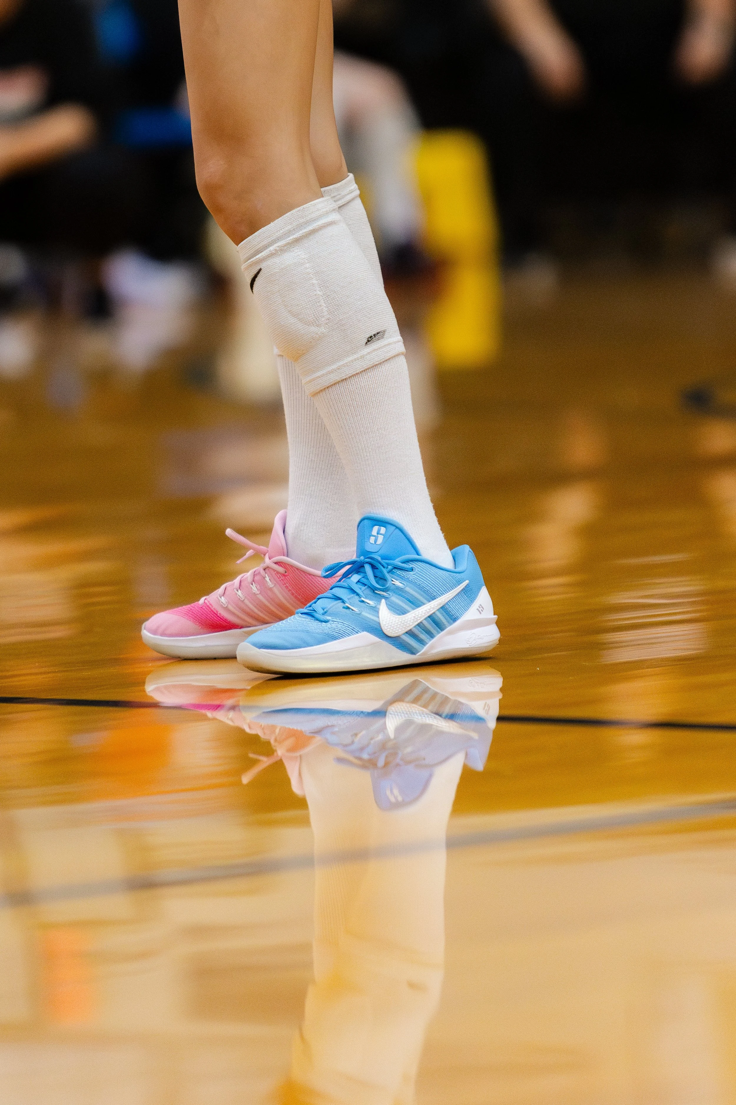 Close-up of a basketball player's feet wearing pink and blue Nike sneakers on a polished wooden basketball court, with reflections visible on the floor.