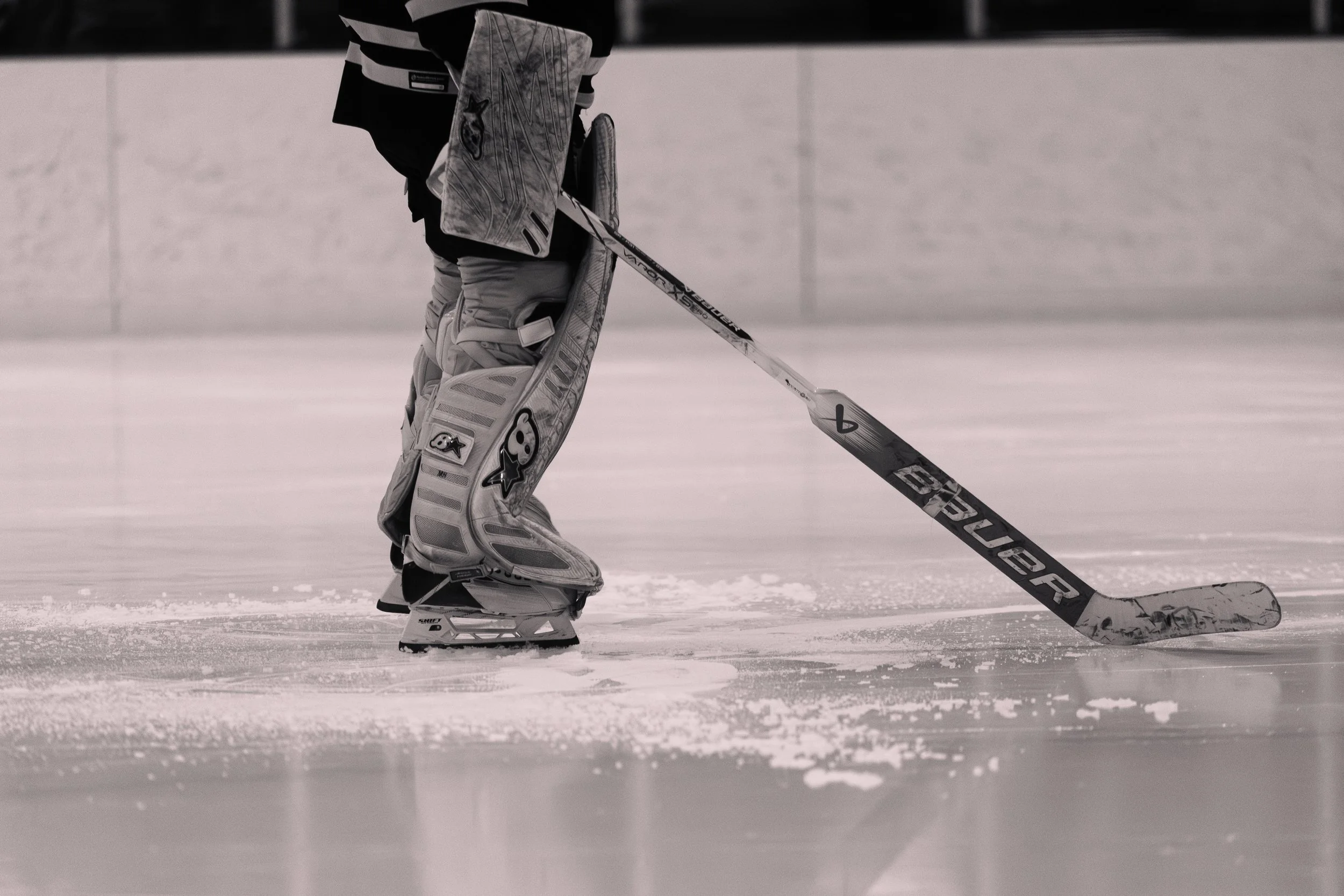 A hockey goalie on the ice in full gear, holding a goalie stick, with a focus on their skates and lower body.