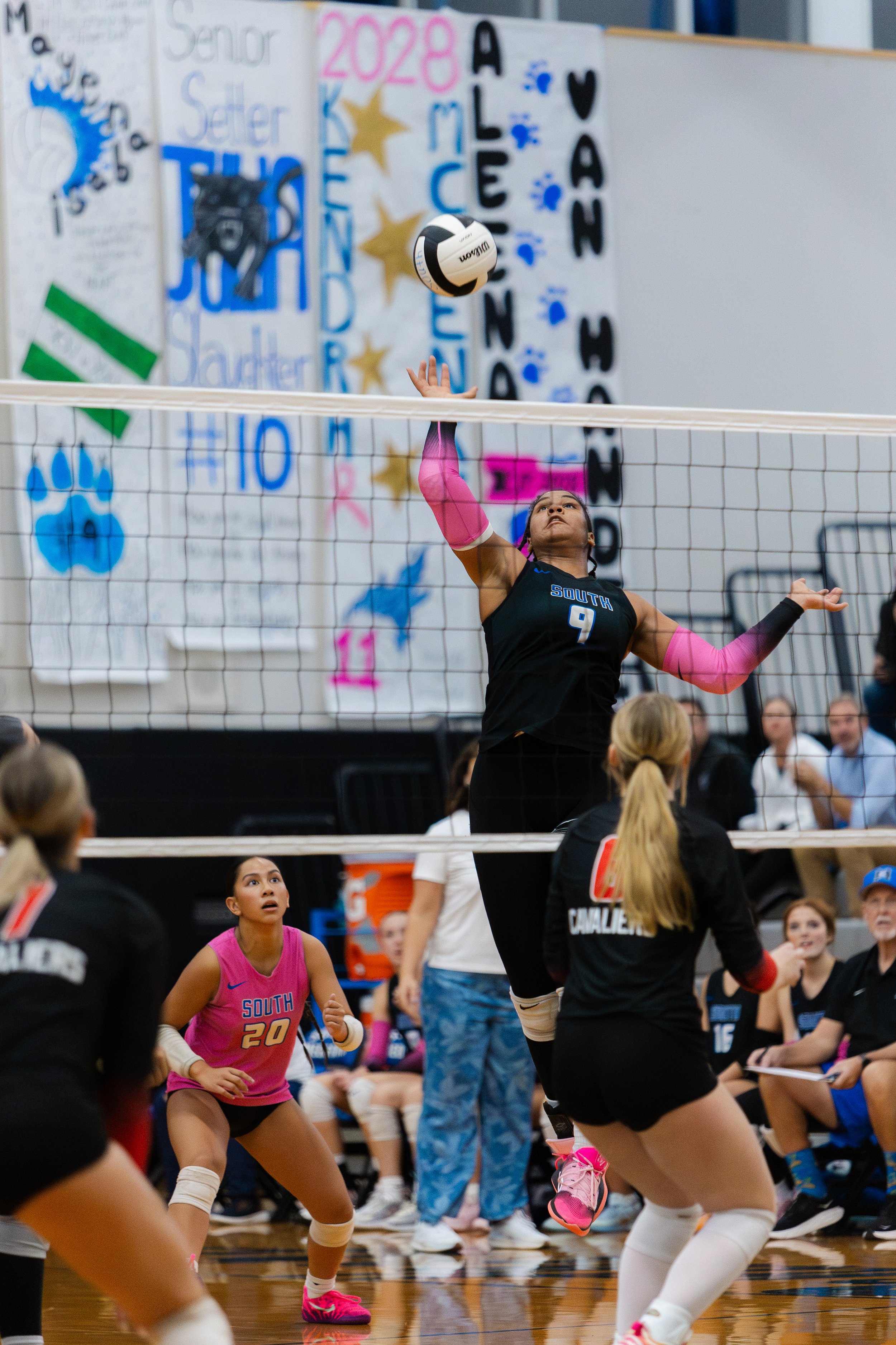 A volleyball player with the number 9 on her jersey, wearing black and pink, jumps to hit the ball over the net during a game. Another player with the number 20 in a pink uniform watches from the ground. Spectators and team members are seated in the 
