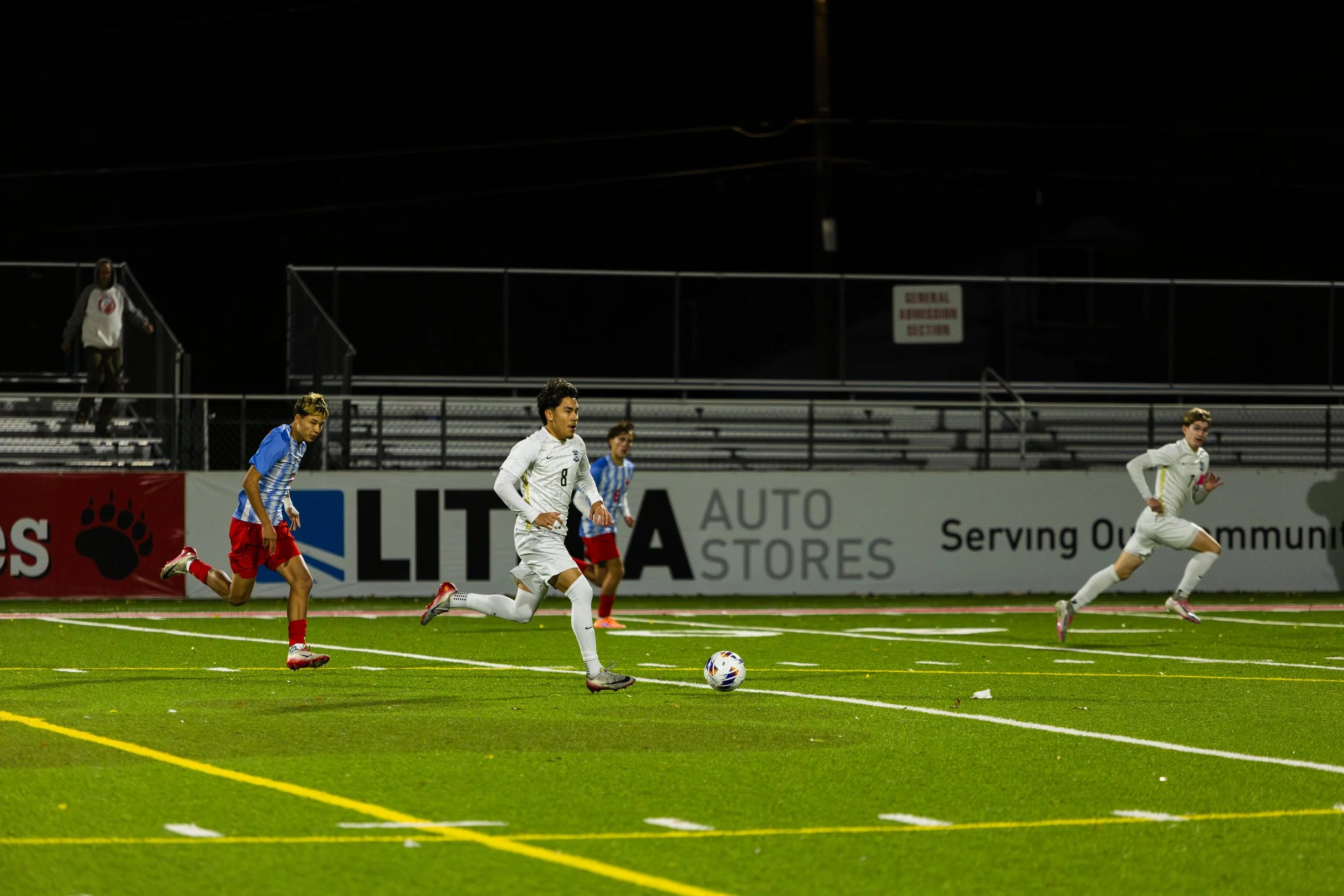 Soccer players running on a field at night, with one player in white controlling the ball, while others in white and blue uniforms chase behind. Empty bleachers and advertising banners are in the background.