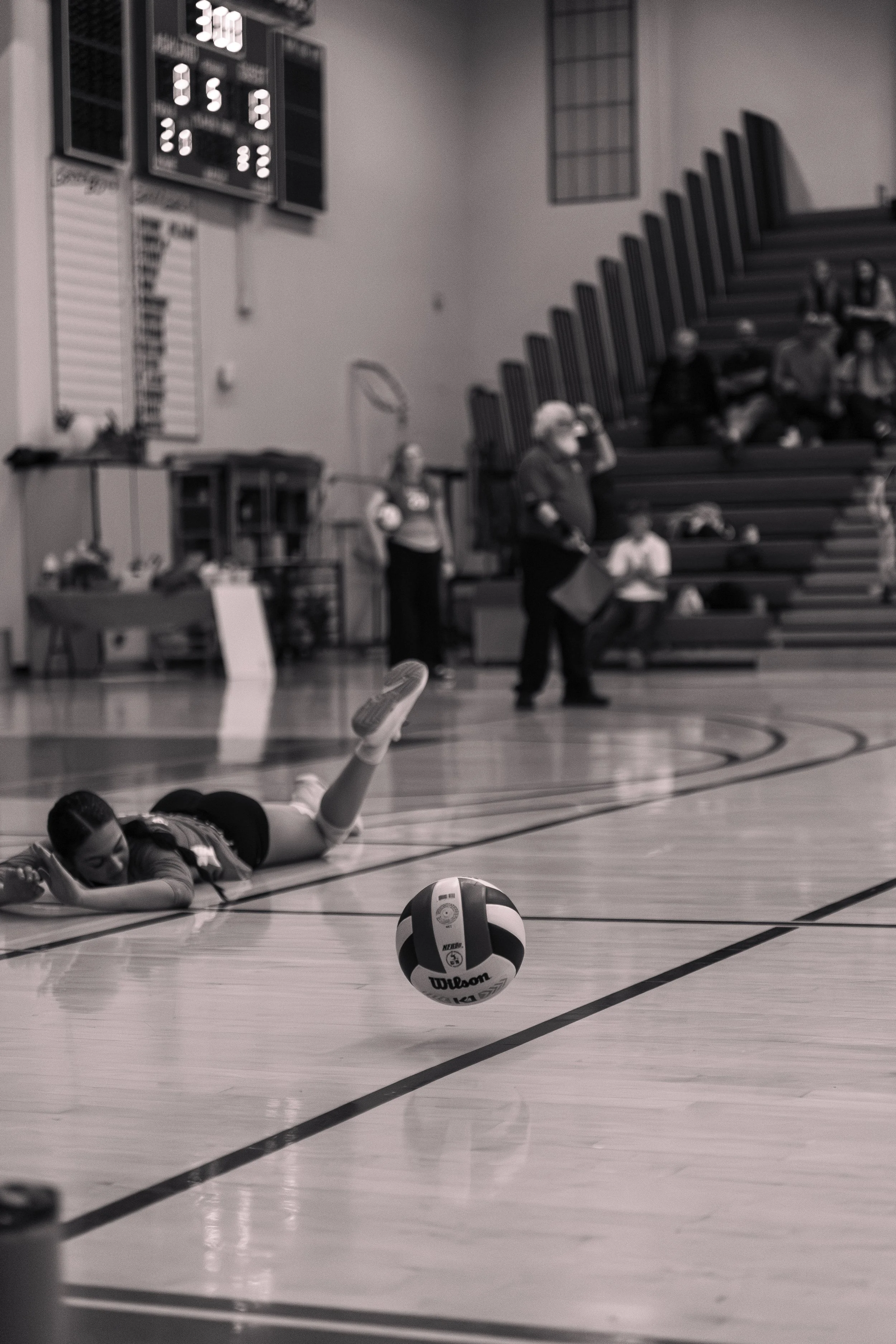 A girl lying on the gymnasium floor during a volleyball game in a gym, with a volleyball ball near her, and spectators sitting on the bleachers in the background.