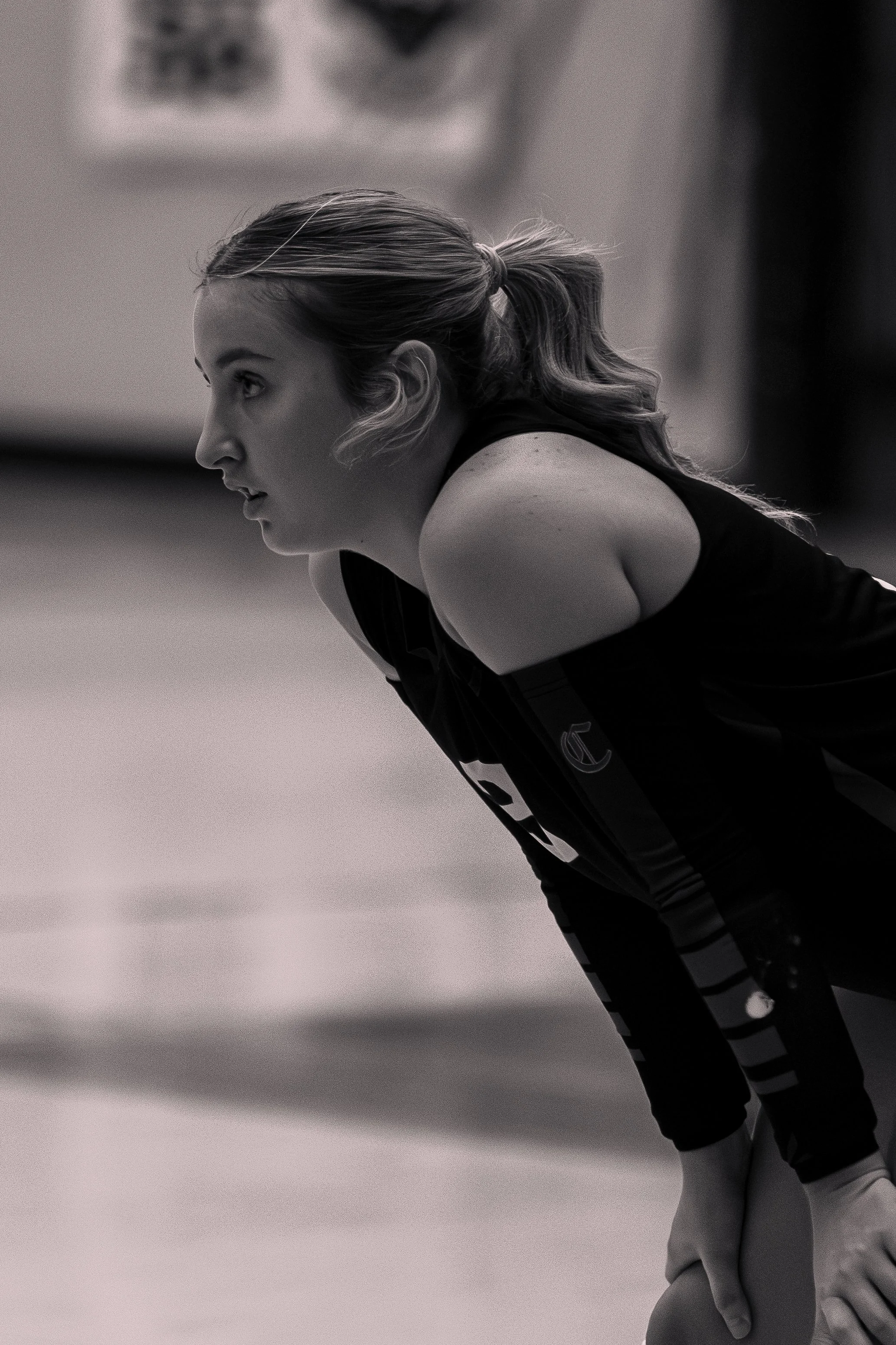 A black and white photo of a female athlete in a sports facility, bending forward with hands on her knees, appearing tired or concentrated.
