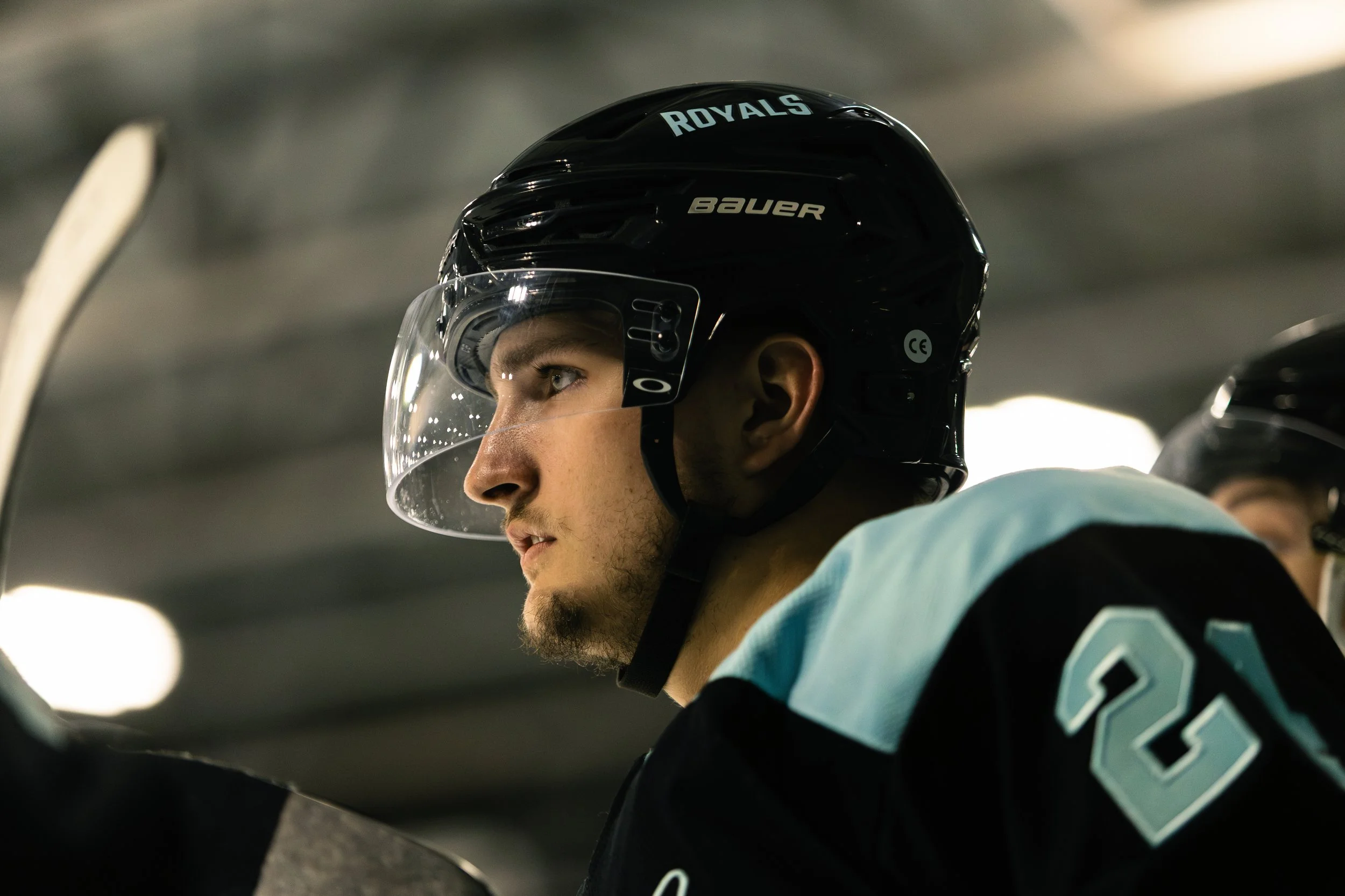 Close-up of a young male hockey player wearing a black helmet with the word 'ROYALS' and a clear visor, sitting on the bench in an indoor ice rink.