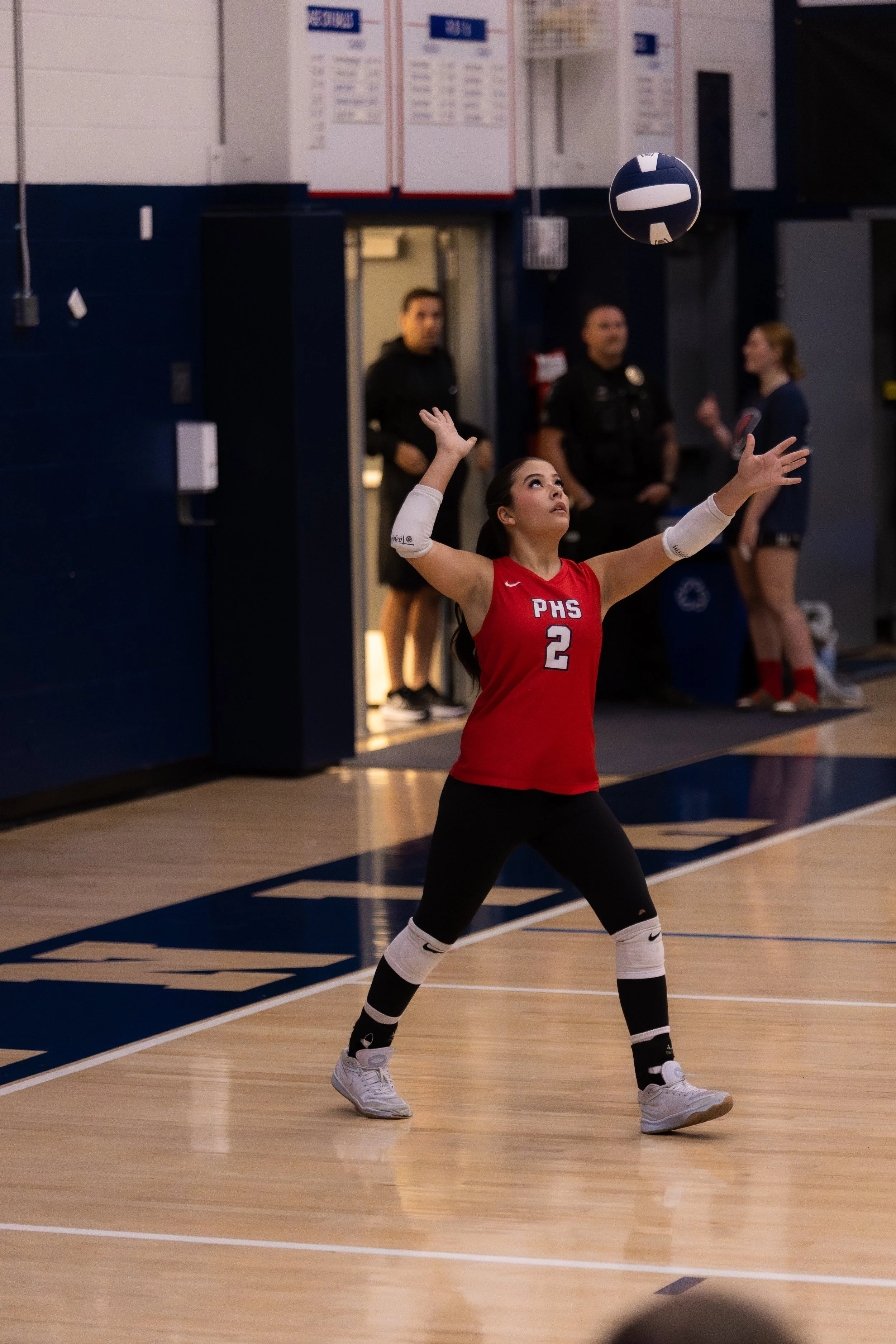 A female volleyball player in a red jersey with the number 2 is preparing to receive the ball during a game.