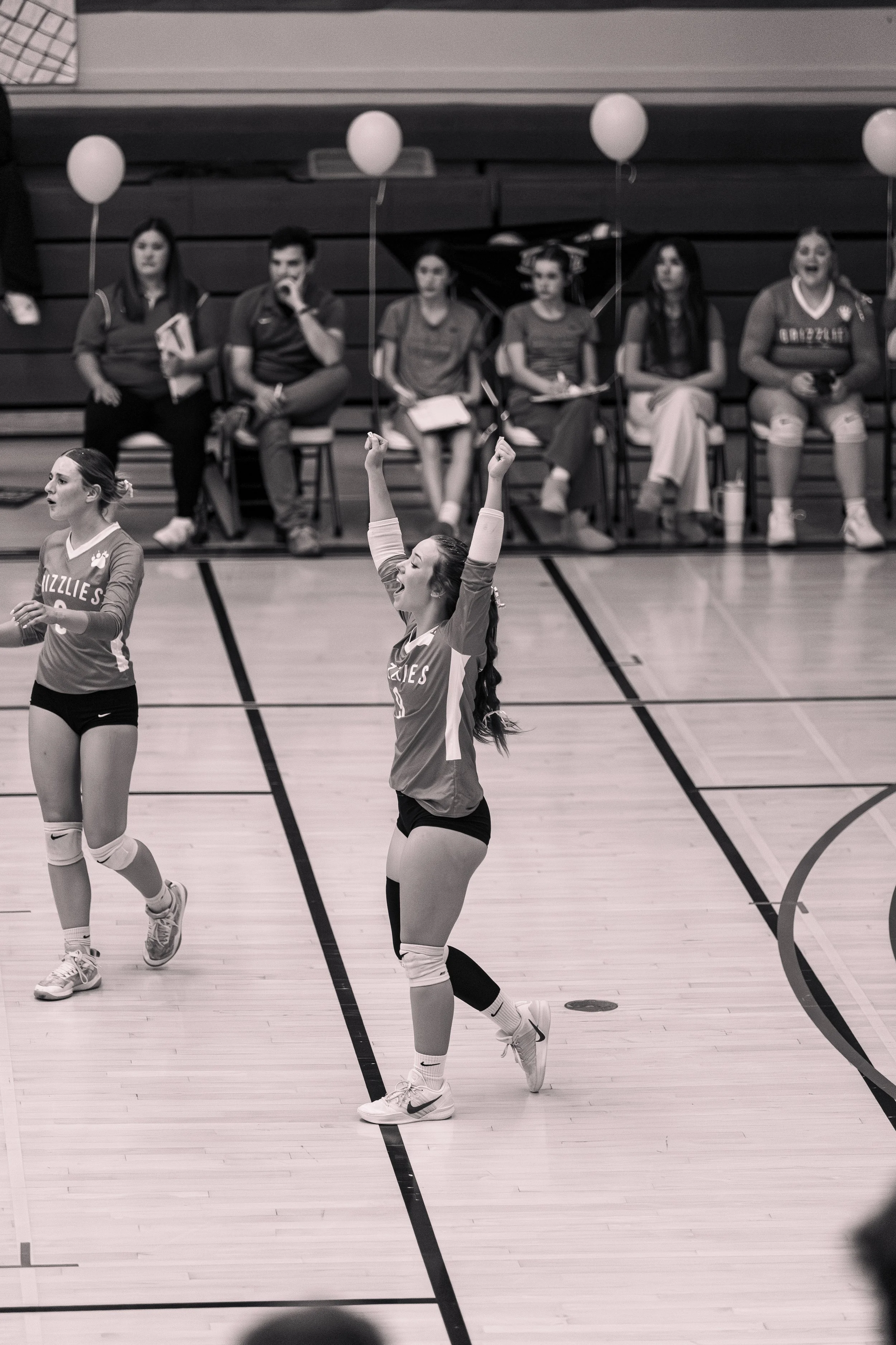 A volleyball player in a jersey with 'Grizzlies' on it celebrates on a gymnasium court, raising both arms in victory. In the background, other players and spectators sit on benches, some with balloons.