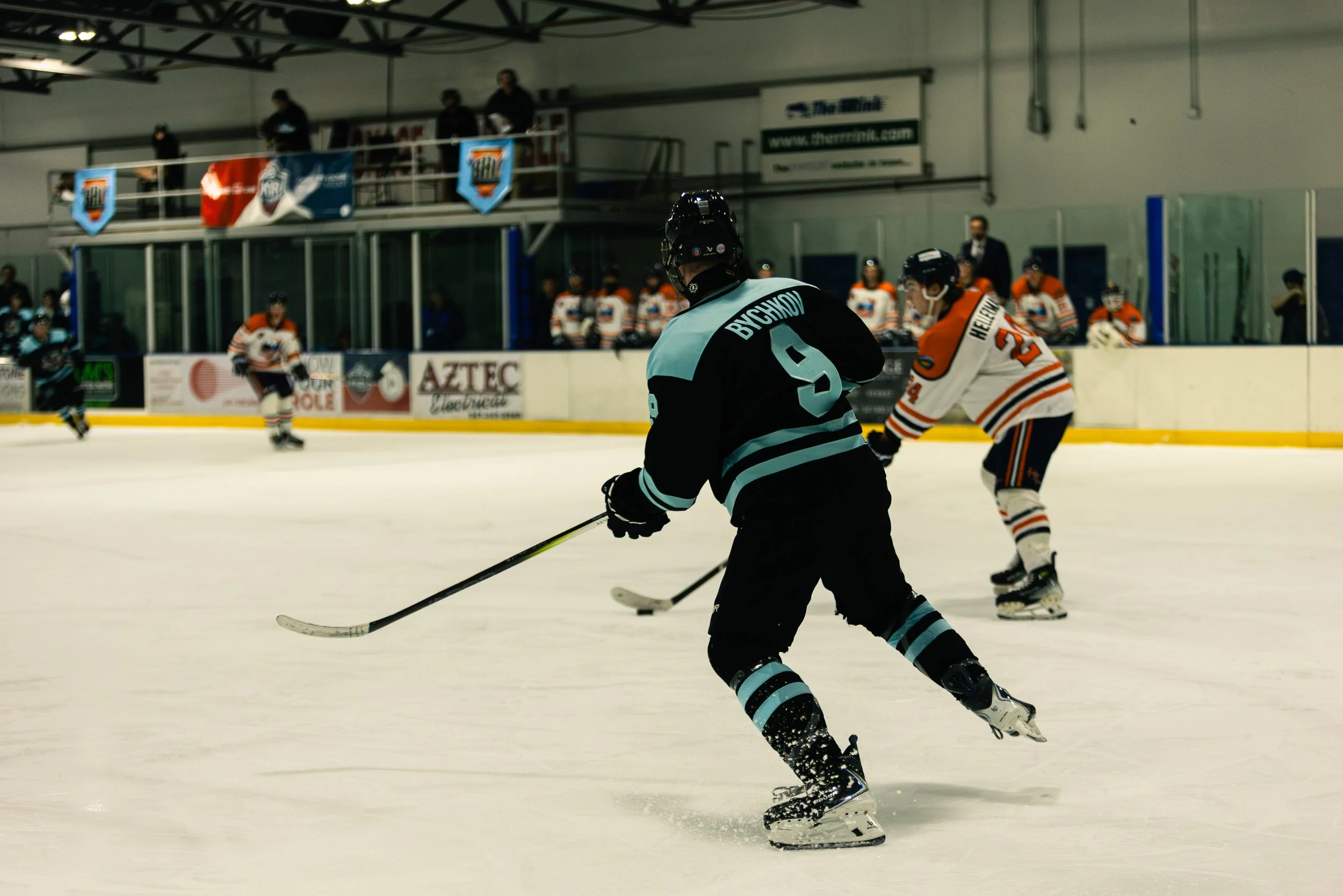 Hockey players in action during a game at an indoor ice rink, with one player in black and teal jersey skating with the puck, and another player in white and orange jersey nearby. Players on the bench are visible in the background.