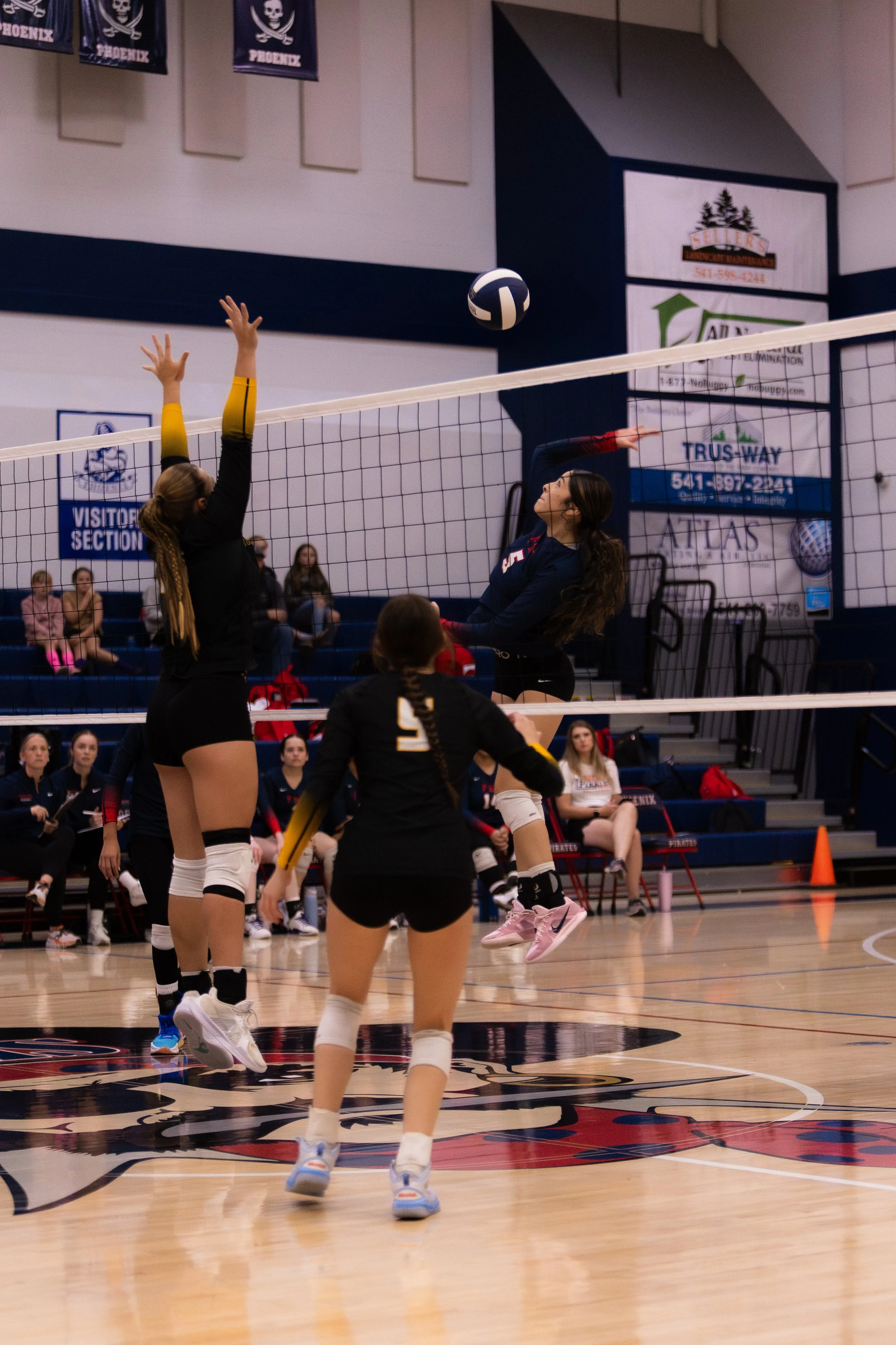 A volleyball game in progress with players at the net, one jumping to hit the ball and another attempting to block. Spectators are seated in the background on bleachers.