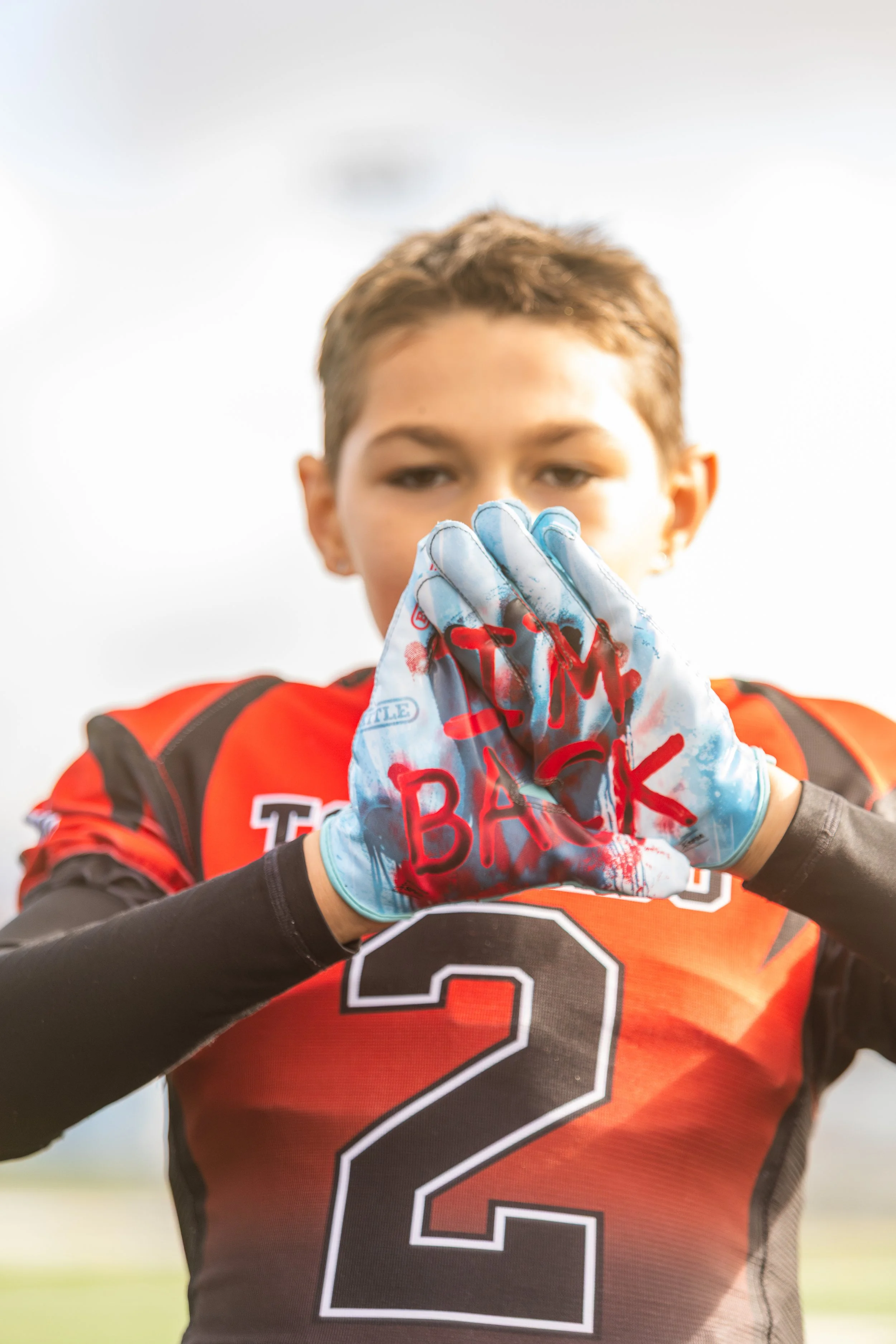 A young football player in a red and black jersey with the number 2, holding his custom painted glove with the words 'TEAM BACK' written in red on his gloves, standing outdoors.