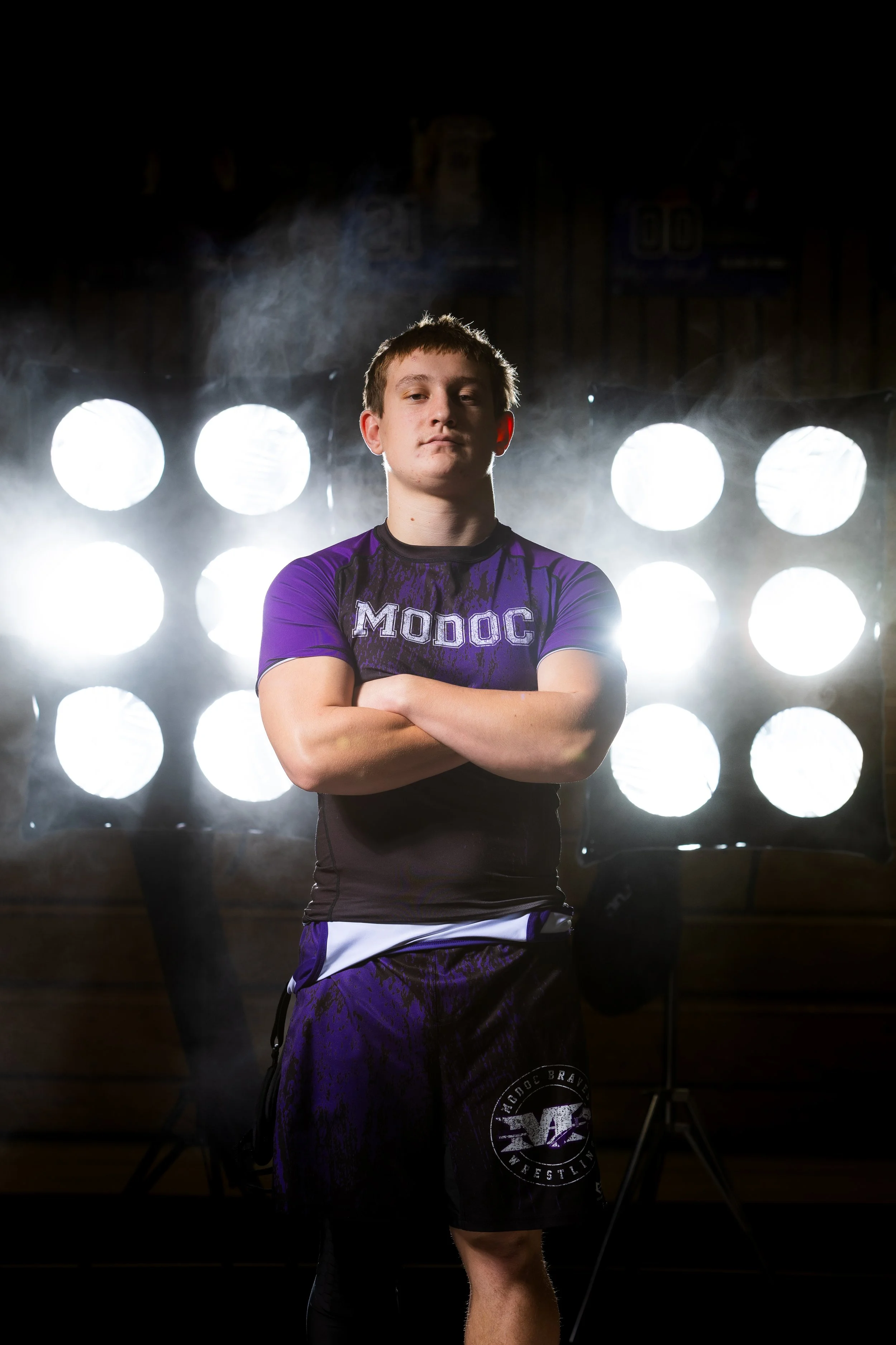 Young male wrestler standing with crossed arms, wearing a purple and black wrestling uniform with 'MODOC' on the shirt, in front of a bright light setup with multiple circular lights