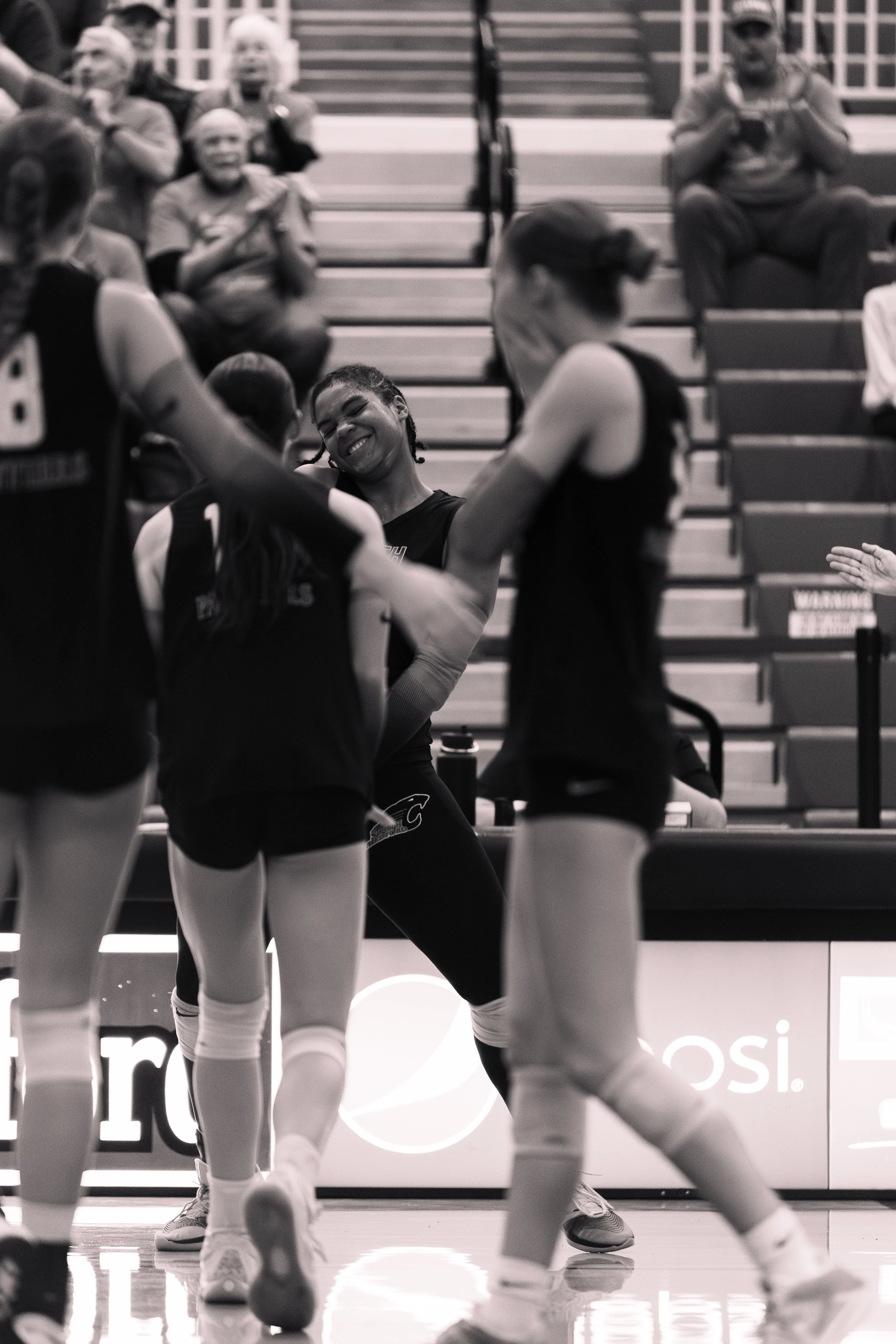 A group of female volleyball players celebrating on the court with some spectators clapping in the background.