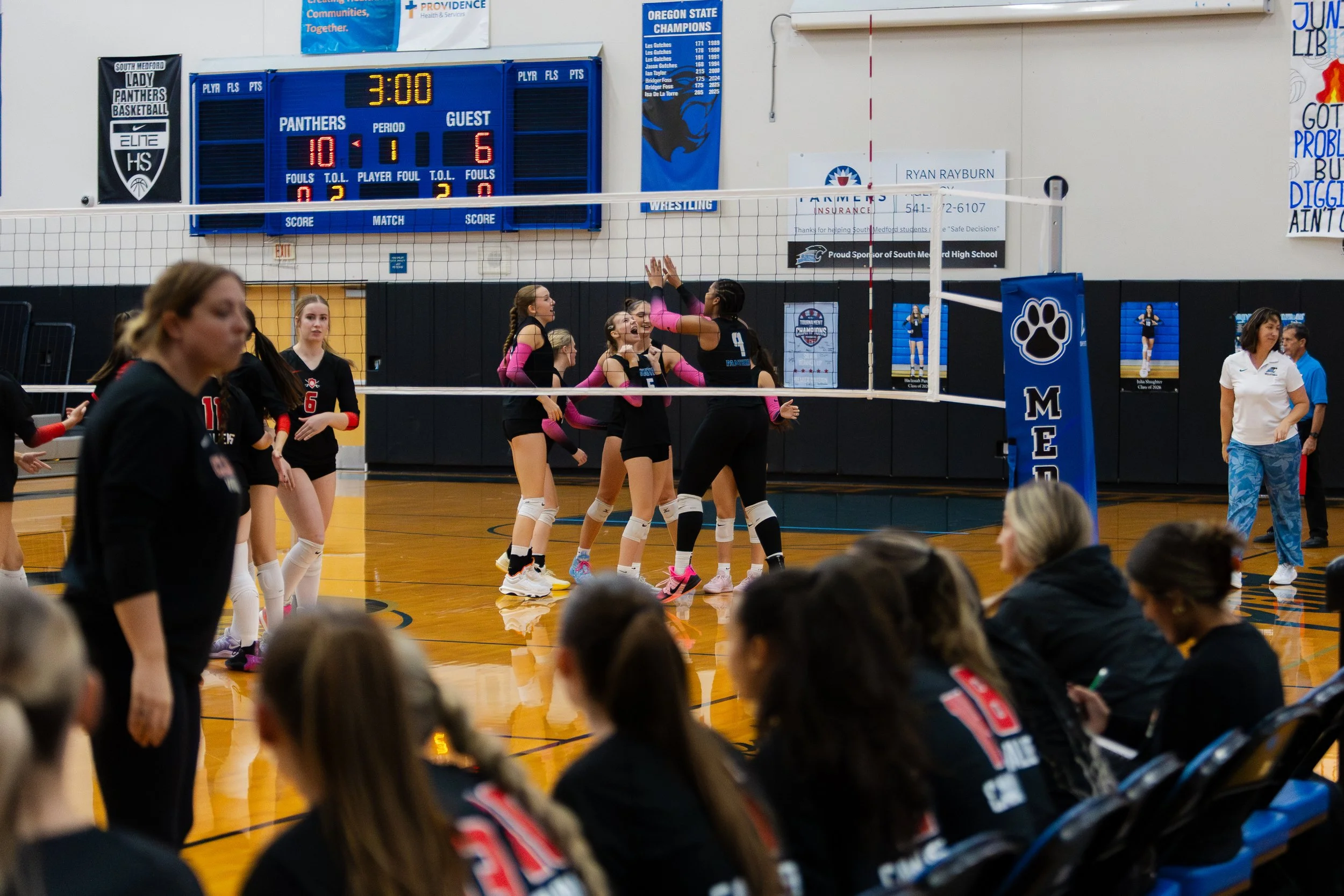 Girls volleyball team celebrating on the court during a match, with the scoreboard showing 10-6 and 3 minutes remaining, in a gymnasium with banners and spectators watching.