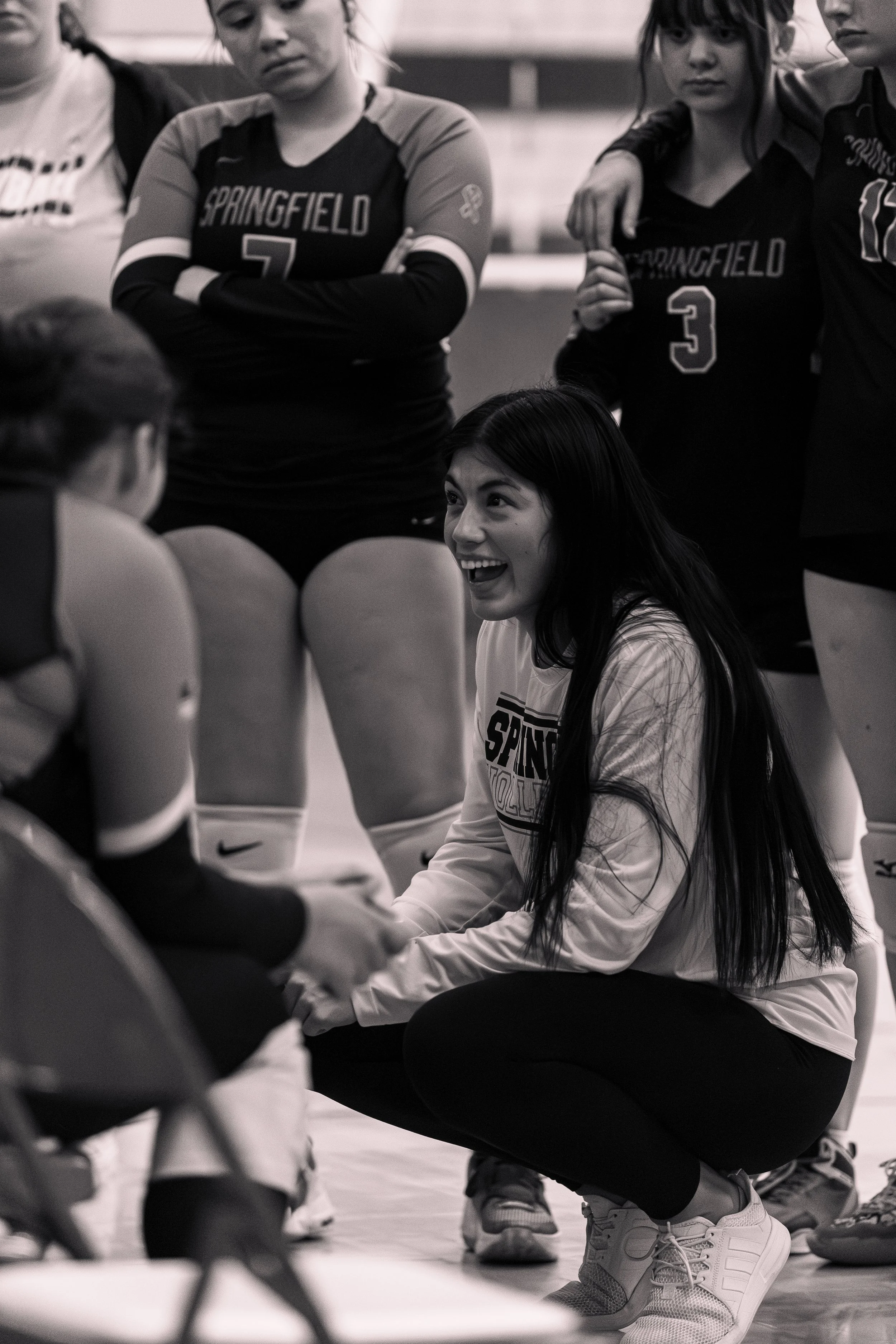 A volleyball coach is kneeling and talking to her team during a timeout, with some players standing and listening.