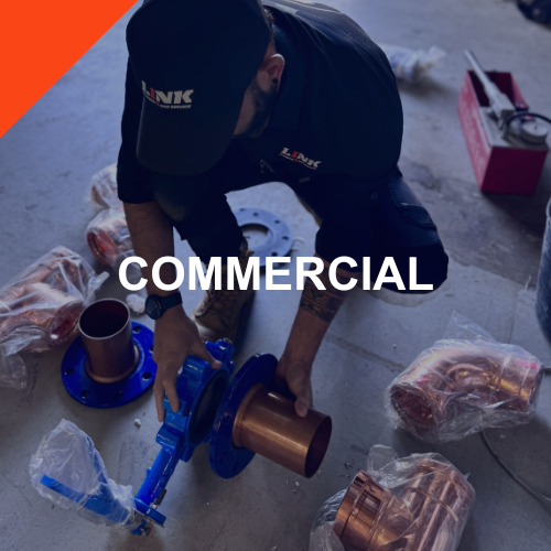 A worker installing plumbing pipes, wearing a black uniform and cap, with the word 'LINK' on it, surrounded by pipes and tools in a construction setting.