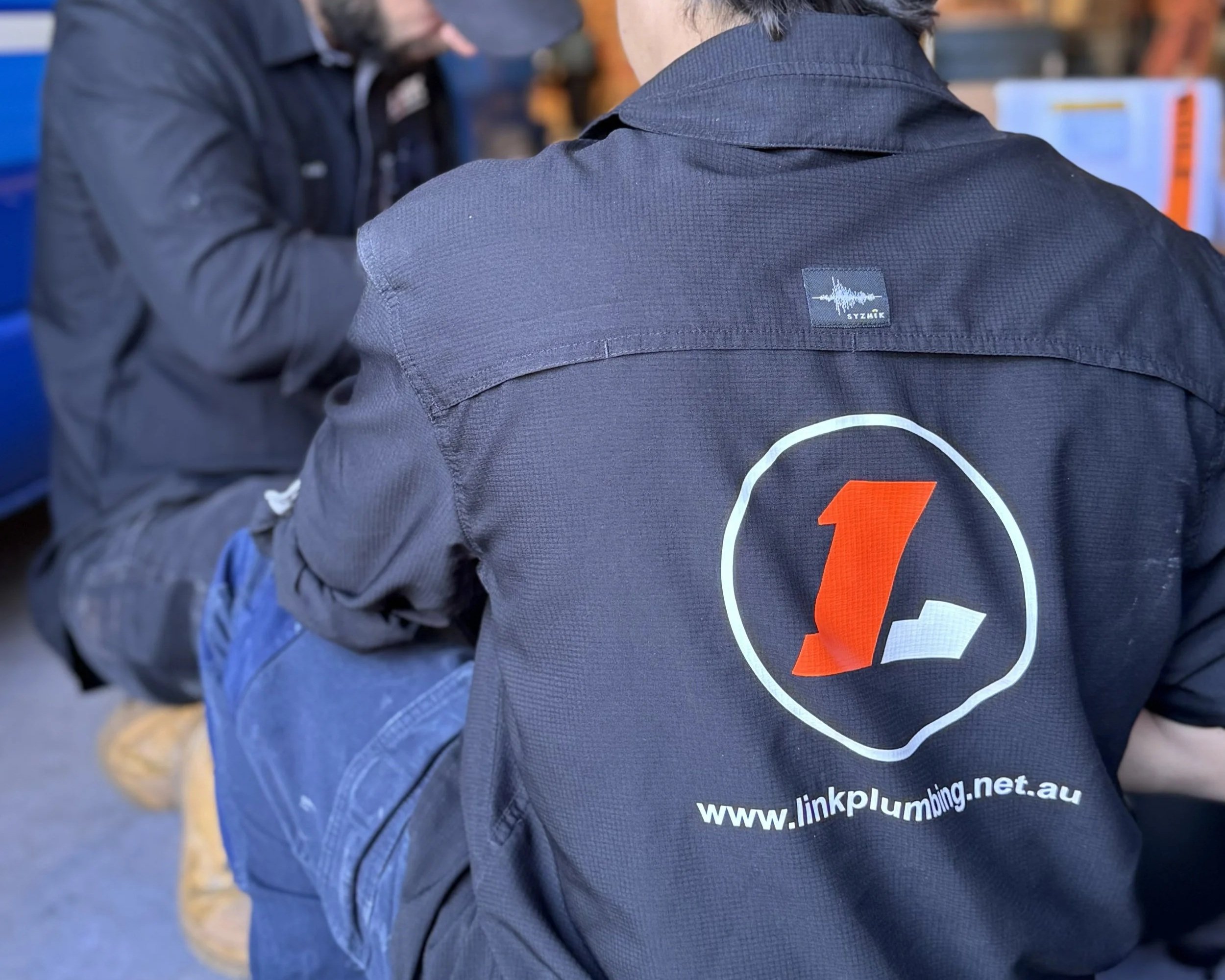 Back of a person wearing a black top with the logo and website of linkplumbing.net.au on it, sitting with another person in the background.