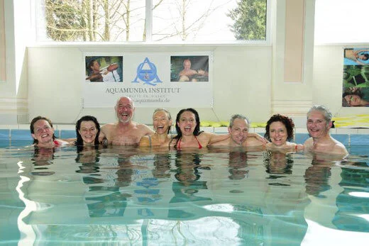 Sieben Menschen, die im Wasser eines Schwimmbeckens stehen, lachen und Spaß haben, im Hintergrund ein Banner des Aquamunda Instituts.