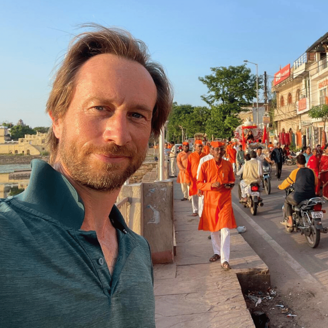 Lincoln Harris taking a selfie on a busy street with orange-clad people and motorbikes, with buildings and trees in the background.