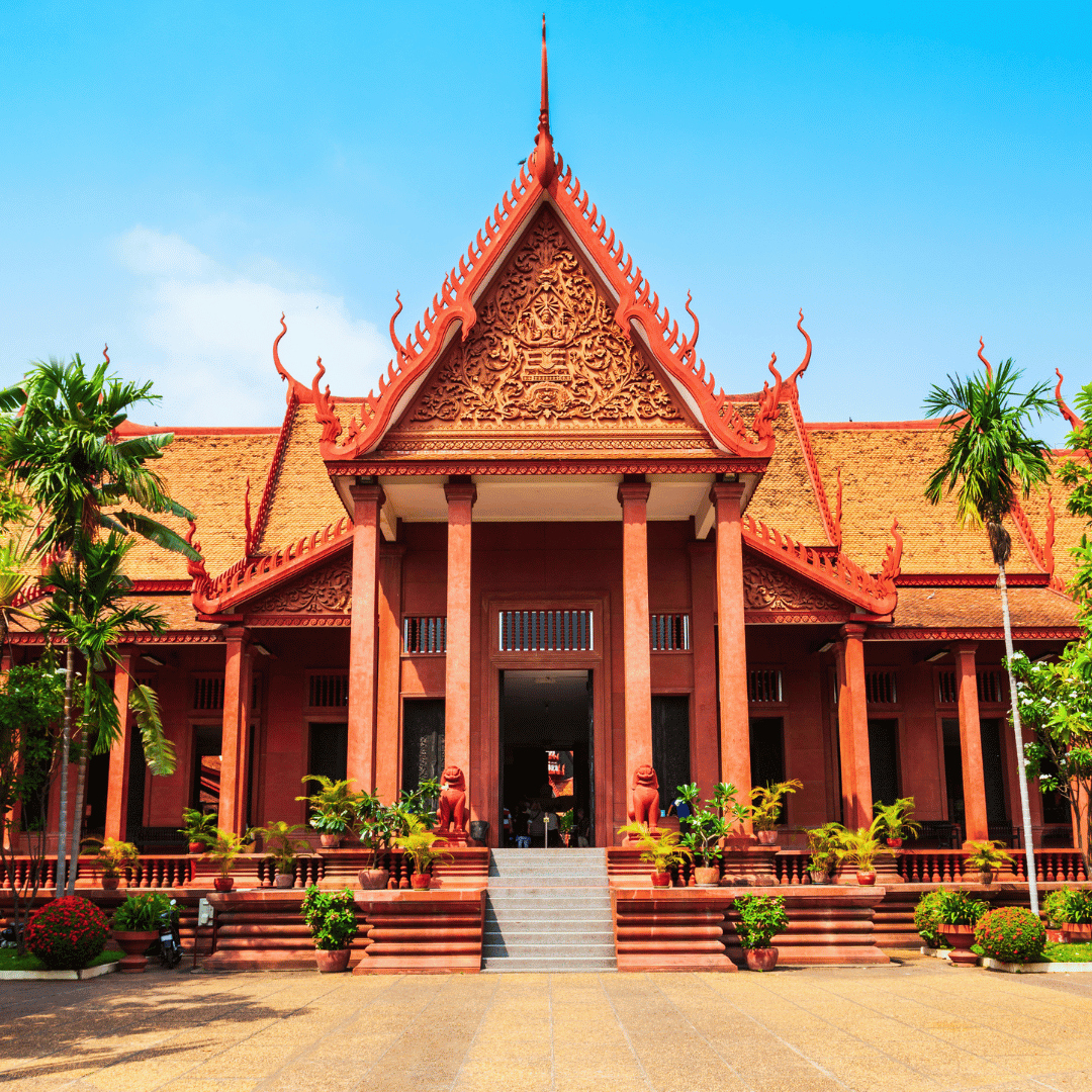 A traditional Thai temple with ornate red architecture, tall pillars, and decorative roofing, surrounded by potted plants and palm trees.