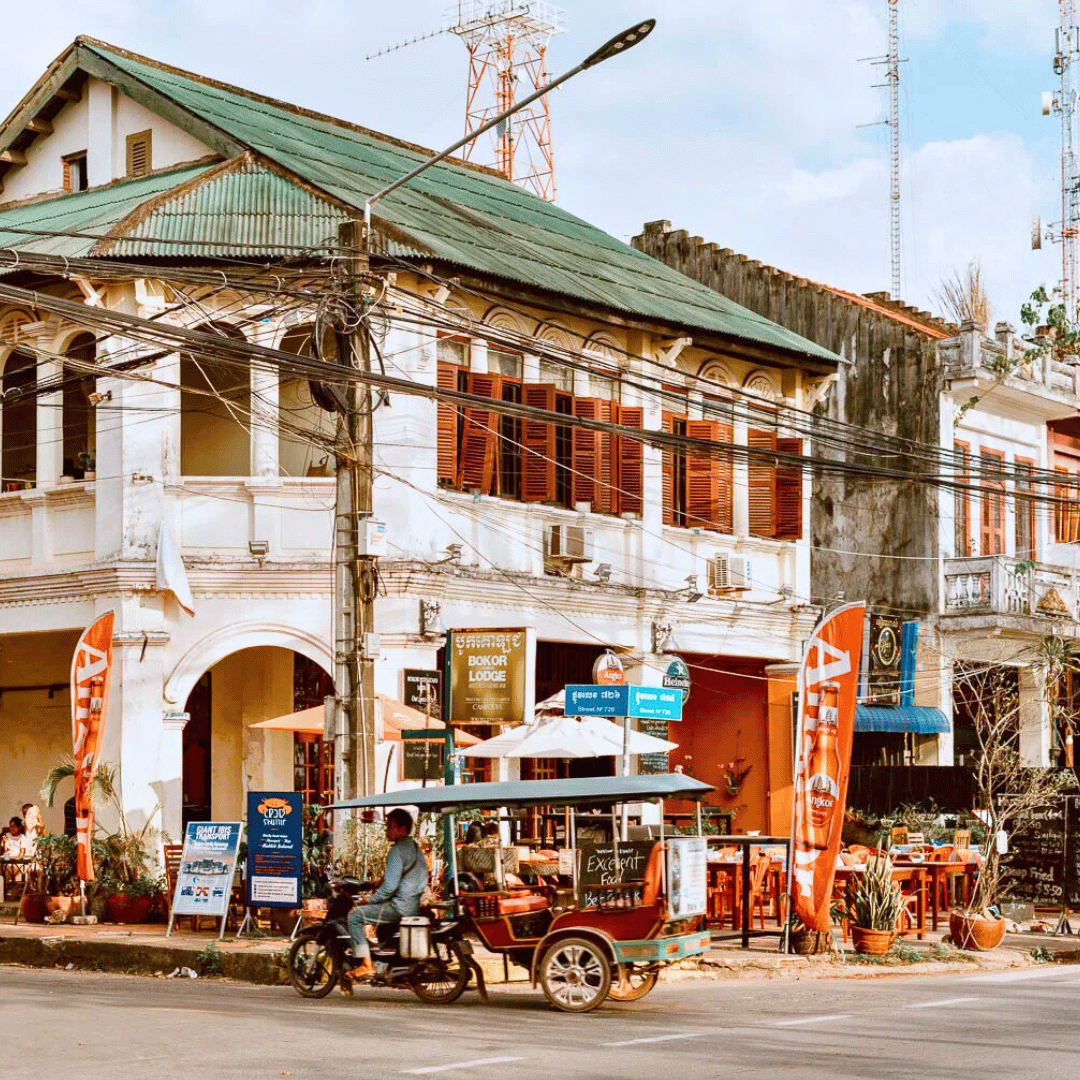 Street view of a two-story building with a green sloped roof, wooden shutters, and a cafe or restaurant on the ground floor. A motorized tuk-tuk vehicle is in front of the building. There are flags, signage, and outdoor seating with umbrellas and potted plants.