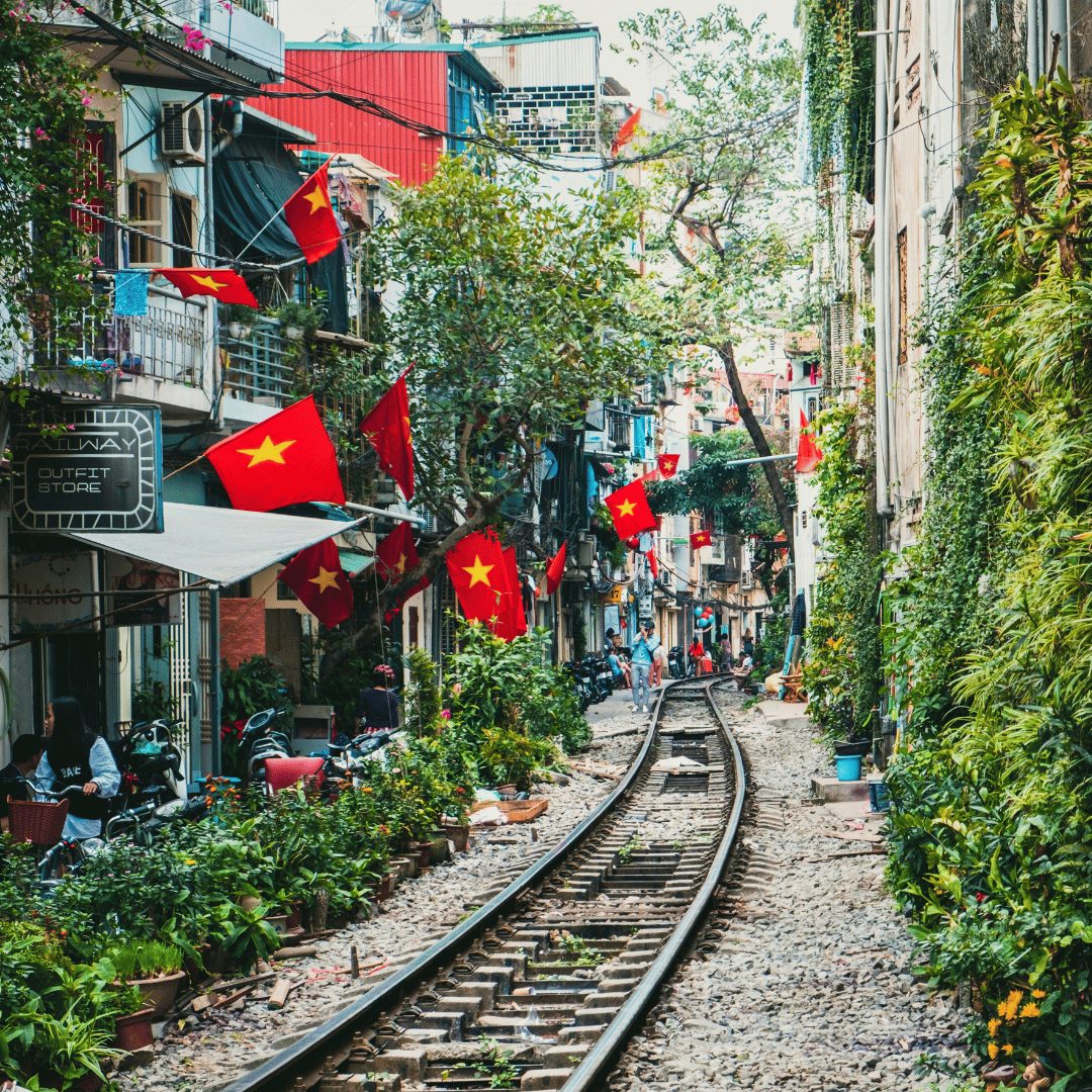 A narrow urban street with train tracks running down the center, decorated with Vietnamese flags. Buildings line both sides with balconies, plants, and shops. People are seen walking and sitting, with greenery and bright colors throughout.