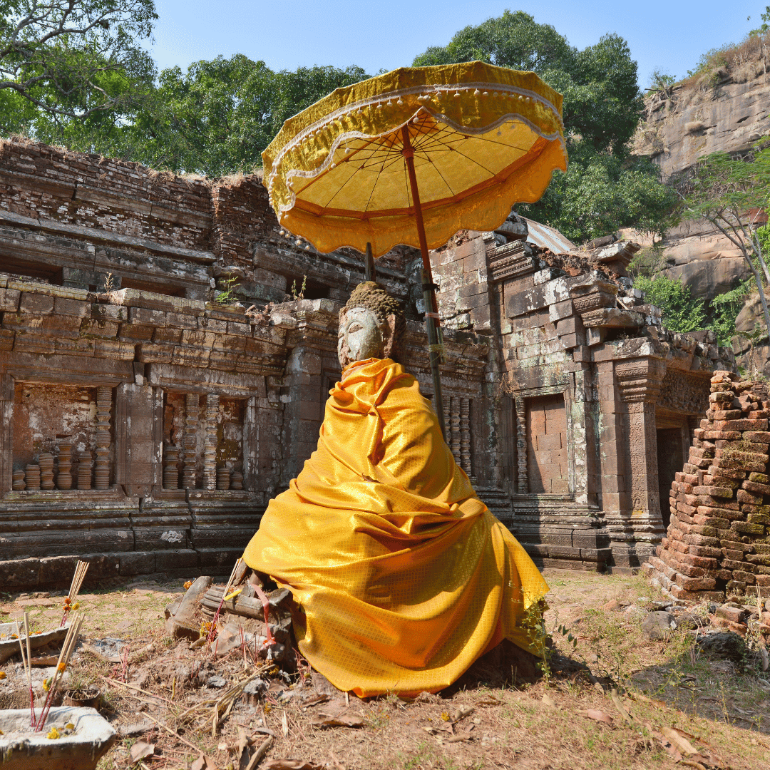 A Buddha statue draped in a yellow cloth, seated outdoors in front of ancient stone ruins, holding a yellow parasol, with green trees and rocky cliffs in the background.