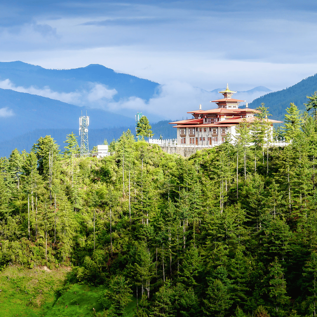 A traditional Asian-style temple with multiple tiers and a pagoda roof, situated on a lush green hillside surrounded by tall pine trees, with mountains and a cloudy sky in the background.