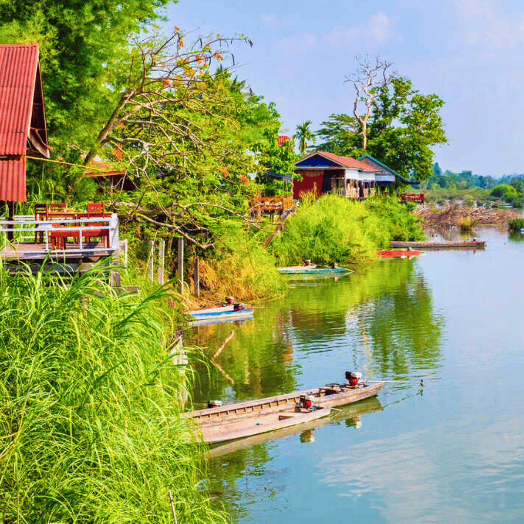 A riverside scene with lush green vegetation, houses on stilts, and boats docked along the riverbank.