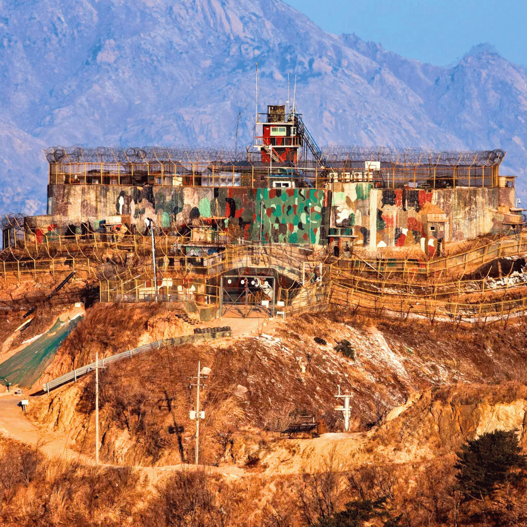 A military bunker with camouflage paint on the walls, surrounded by barbed wire on a hill with dry grass, with mountains in the background.