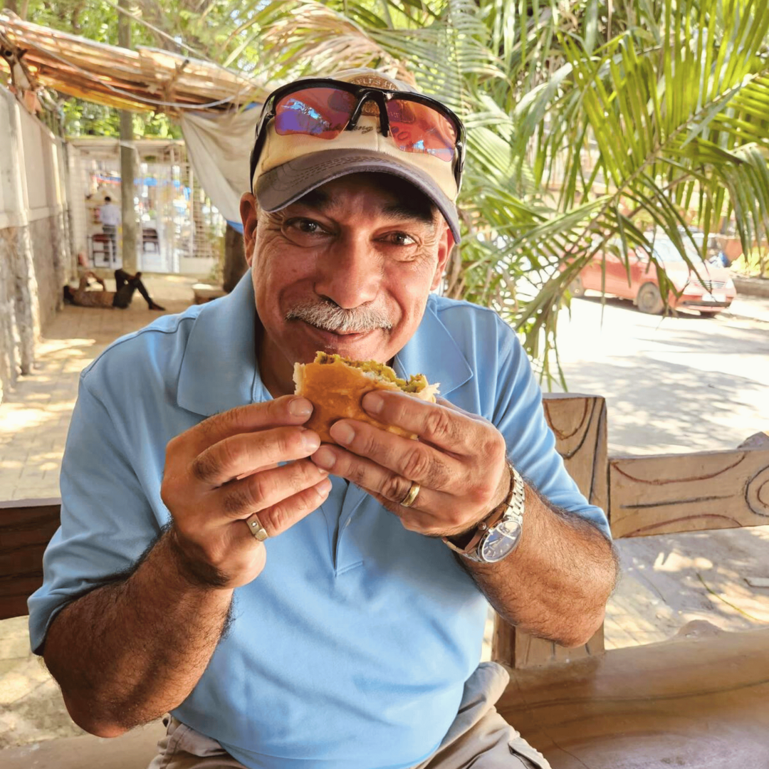 Merrick Abraham in a light blue polo shirt and a beige baseball cap with sunglasses on top, enjoying a sandwich at an outdoor setting with palm trees and a street in the background.