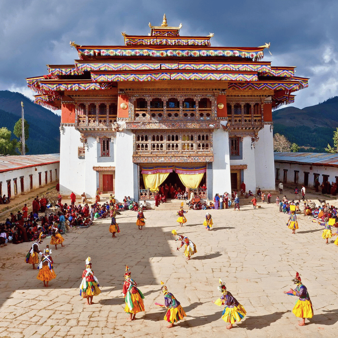 A traditional building with ornate wooden details and colorful decorations, surrounded by people, some dancing in colorful costumes on a stone courtyard, with mountains in the background.