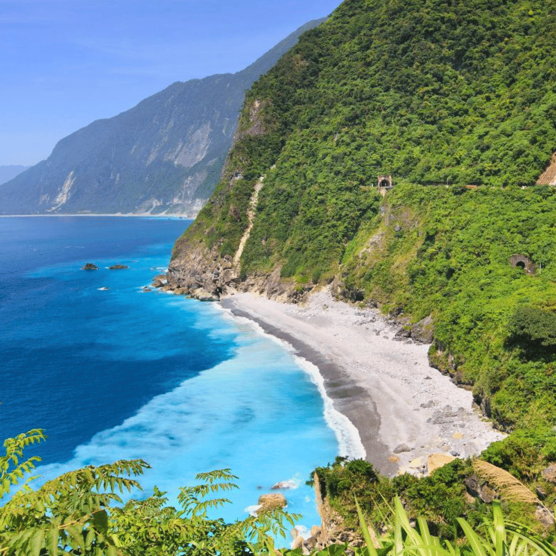 Coastal landscape with a sandy beach, blue ocean waves, steep green hills, and mountains in the background.