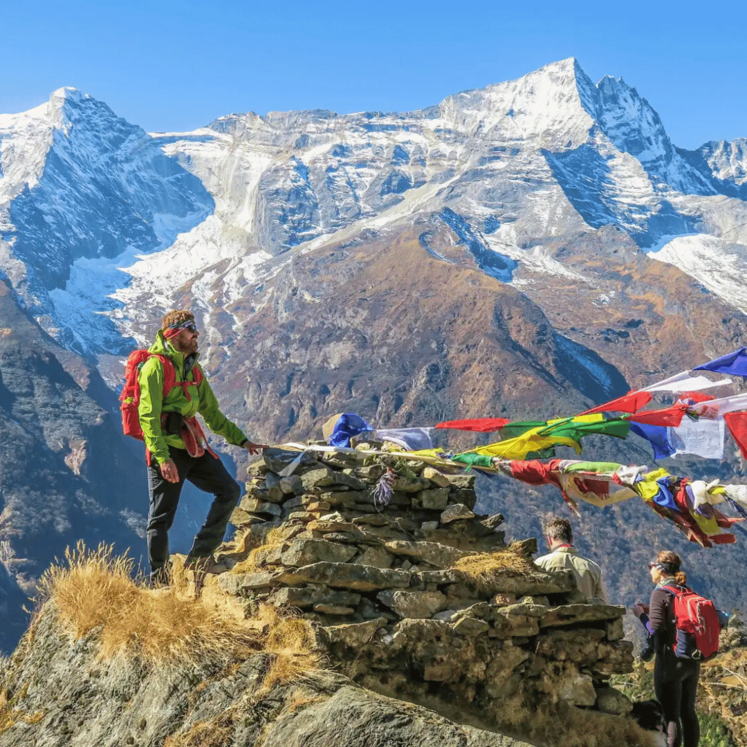 Hikers standing on a mountain trail with a stone cairn and prayer flags, snow-capped peaks in the background.