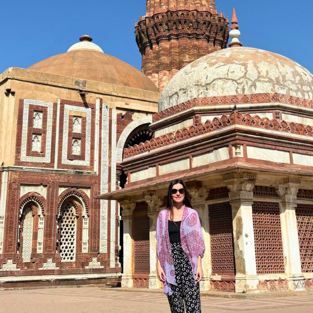 Lorinda Childs standing in front of historic Indian architecture featuring domed structures, intricate red and white stone carvings, and a tall minaret against a clear blue sky.