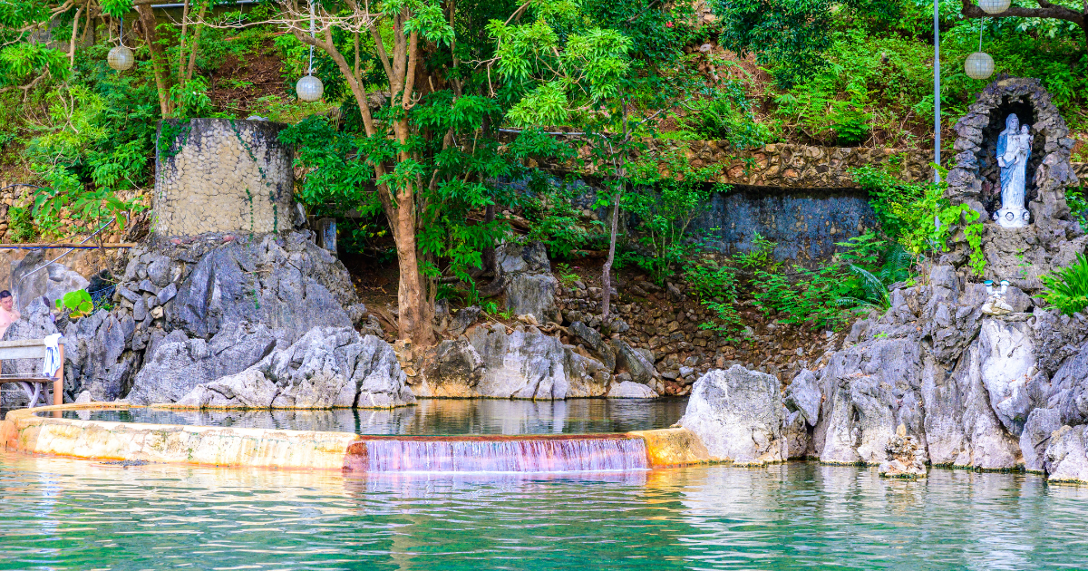 A stone-edged pool at Maquinit Hot Springs with rocks, greenery and a small grotto