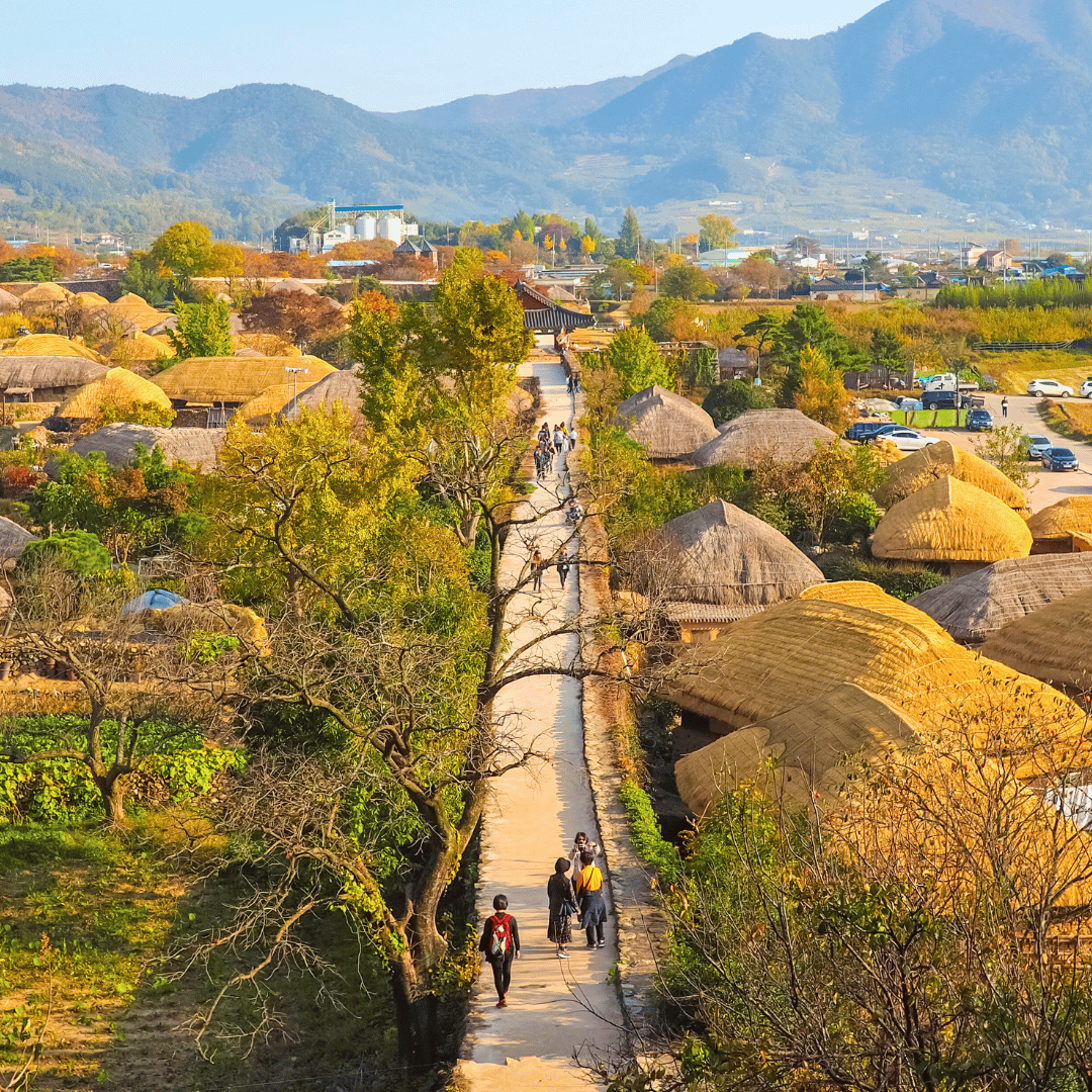 A scenic village with traditional thatched-roof houses, a pathway through colorful trees, and mountains in the background, with people walking along the path.