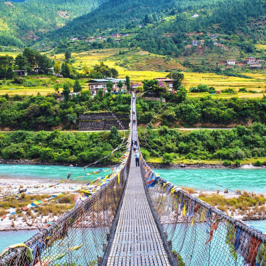 A long suspension bridge decorated with colorful prayer flags stretches across a river with lush green hills and mountains in the background.