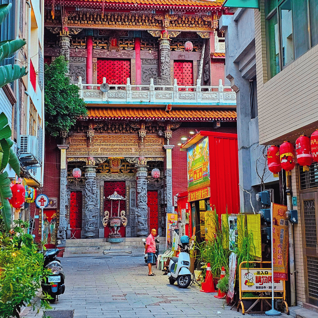 Narrow street leading to a traditional Chinese temple with ornate decor, red doors, and hanging lanterns, surrounded by modern buildings.
