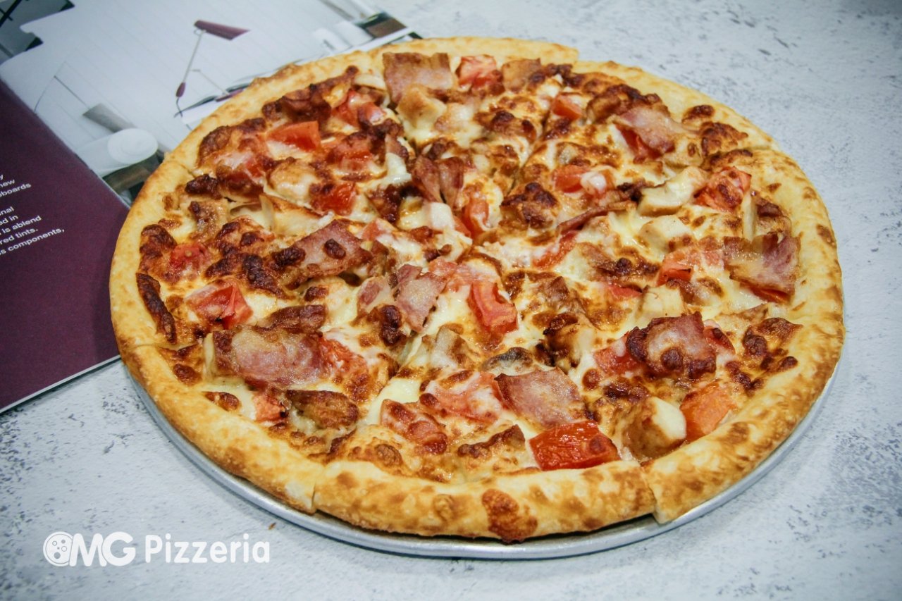 A large pizza with toppings including bacon, tomatoes, and cheese, on a silver tray at a pizzeria.