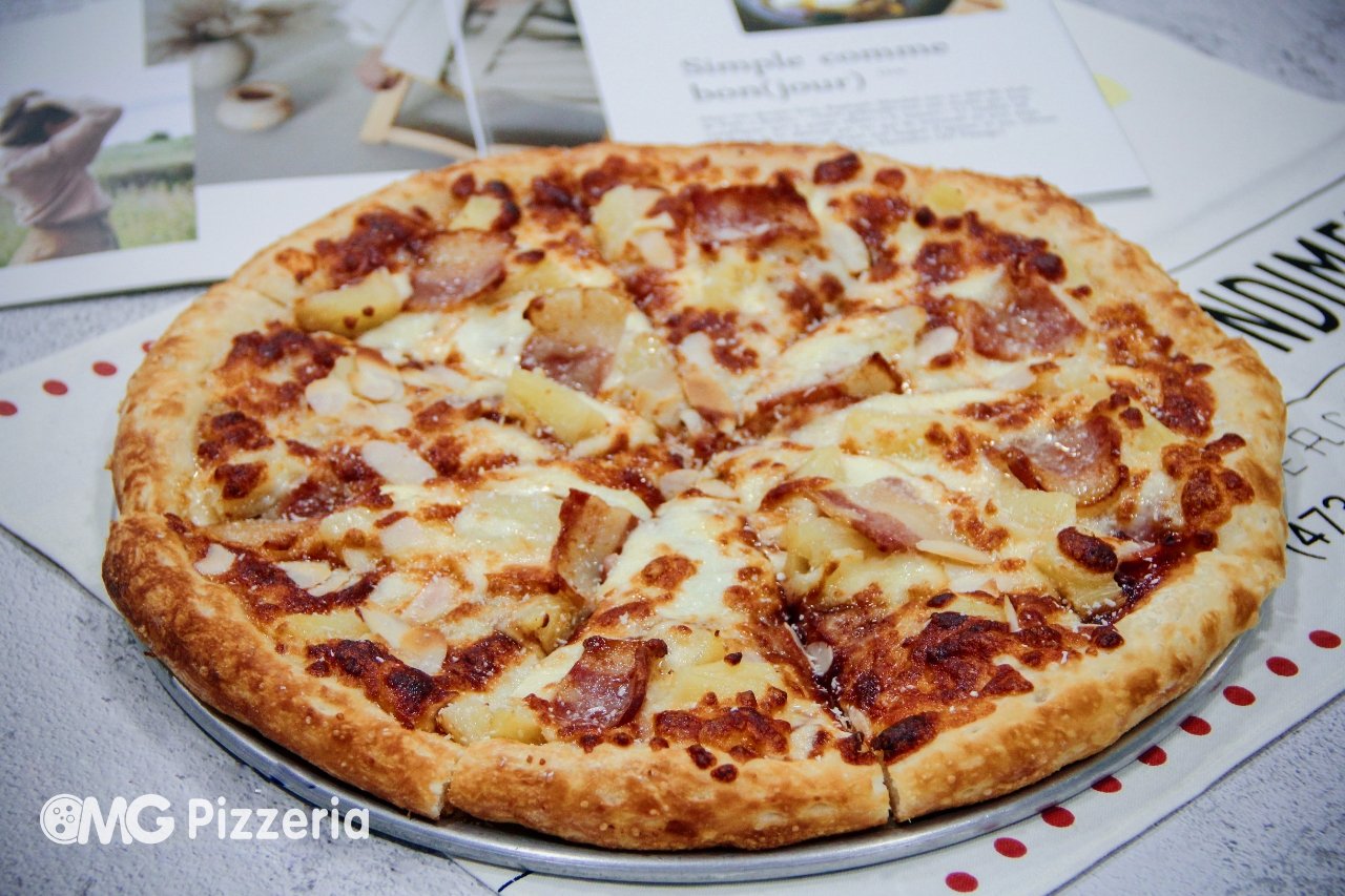 A cheese and ham pizza on a metal tray with a golden crust, topped with melted cheese and chunks of ham and pineapple, placed on a table with papers and photos underneath.