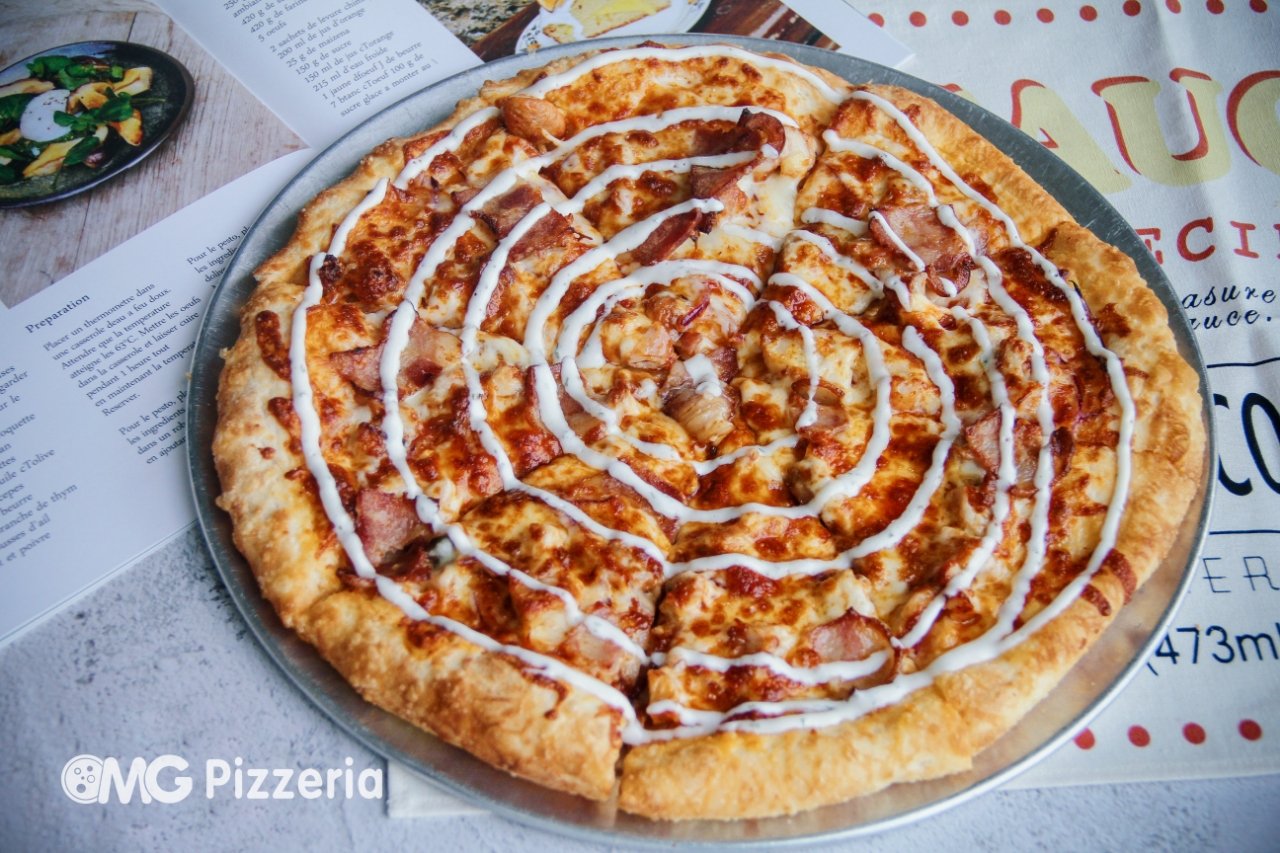 A pizza with a spiral drizzle of white sauce and a base of cheese, ham, and grilled chicken on a metal tray. The tray is on top of a pizza menu and a colorful napkin with red dots.