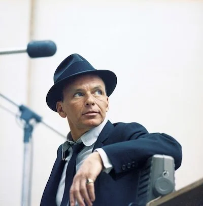 A man in a dark suit and fedora hat sitting next to a microphone, looking to the side.