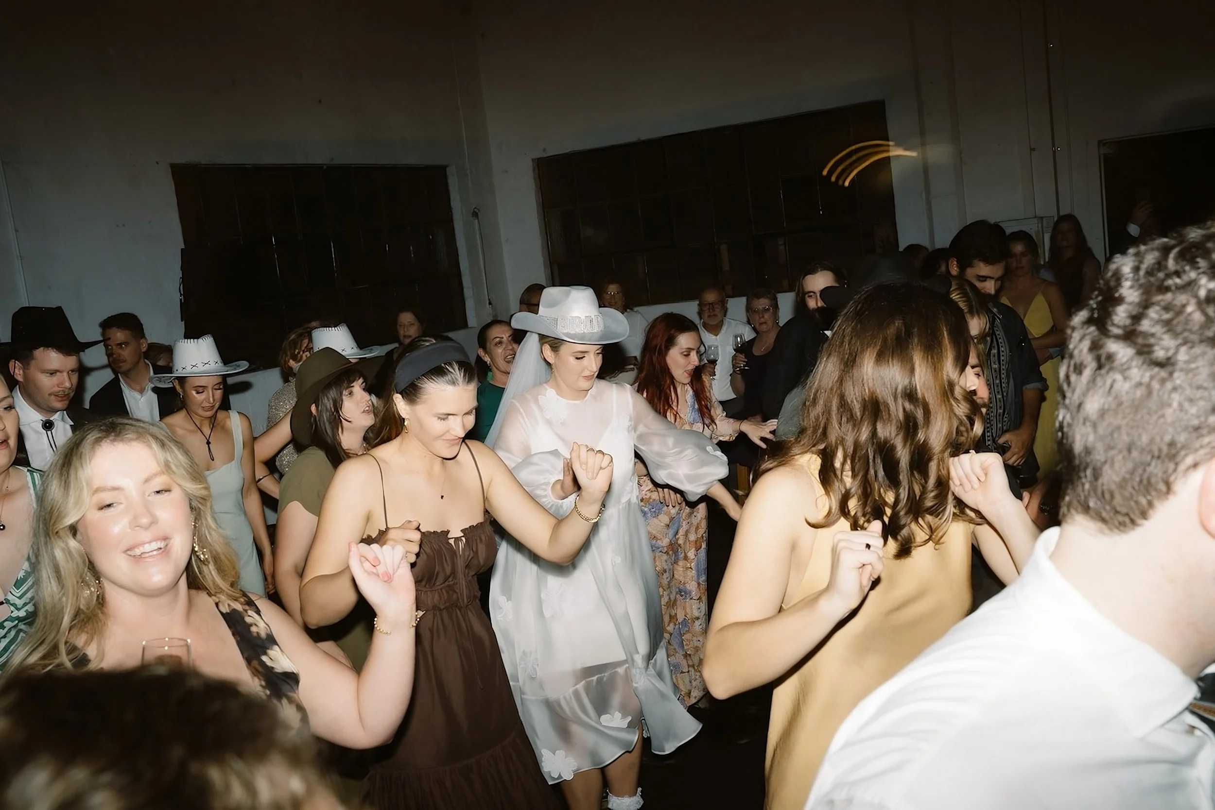 People dancing at a wedding reception, with women wearing hats and bridesmaids, enjoying a lively celebration.
