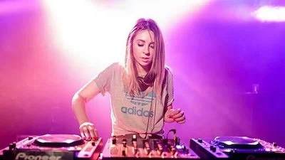 Female DJ with long blonde hair playing music with DJ equipment in a lit club or concert setting with pink and purple lighting.