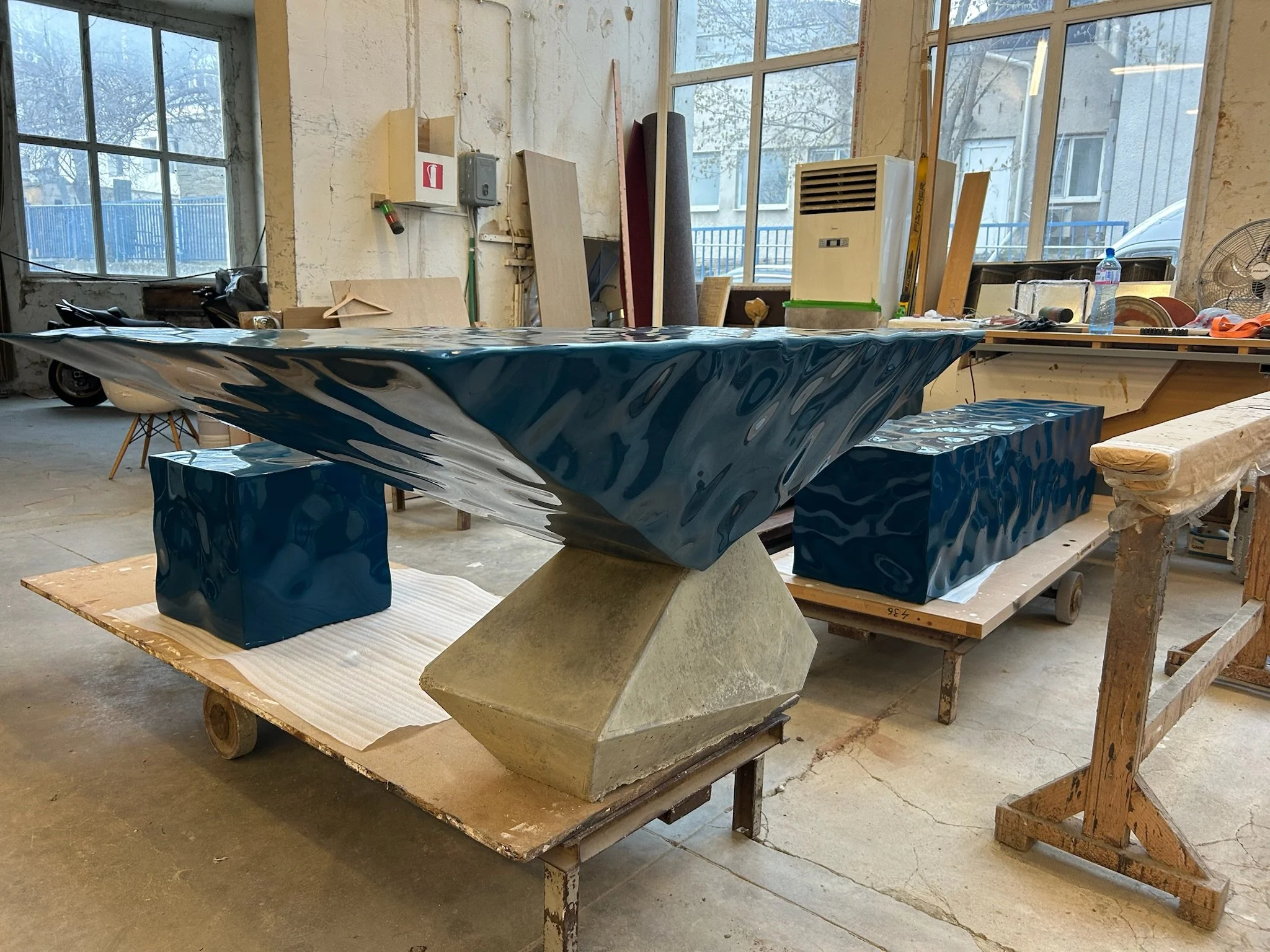 Large blue and black marbled sculpture or artwork resting on a concrete base in a workshop with tools and windows.