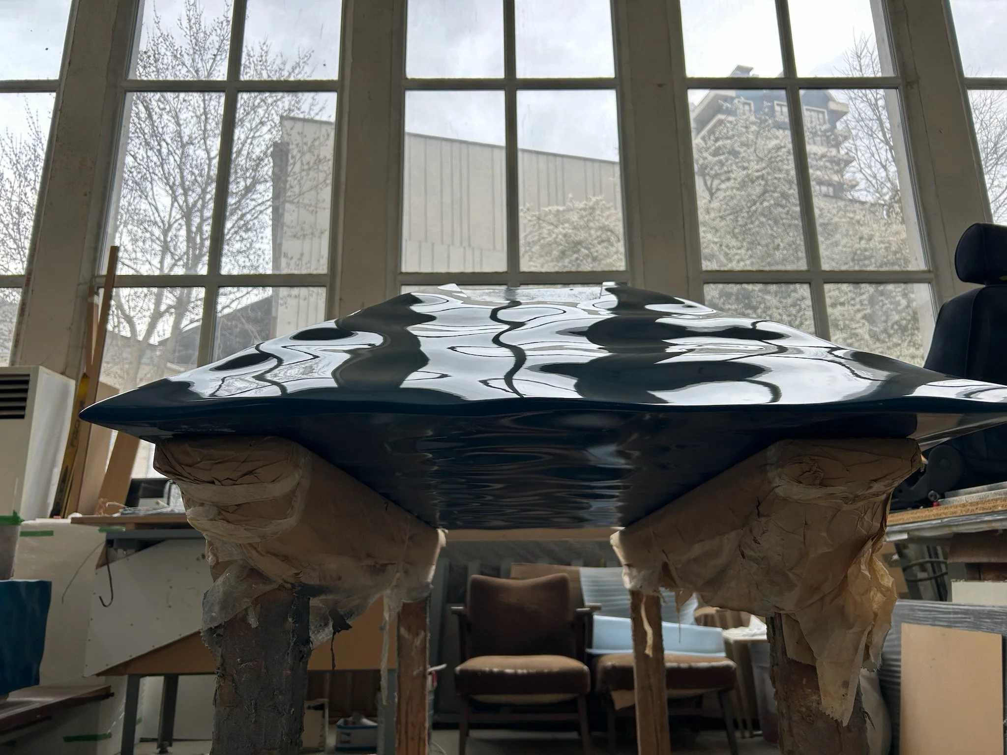 A shiny, glossy blue piece of art, resting on a support structure wrapped in paper in an indoor workshop with large windows and outdoor trees visible.