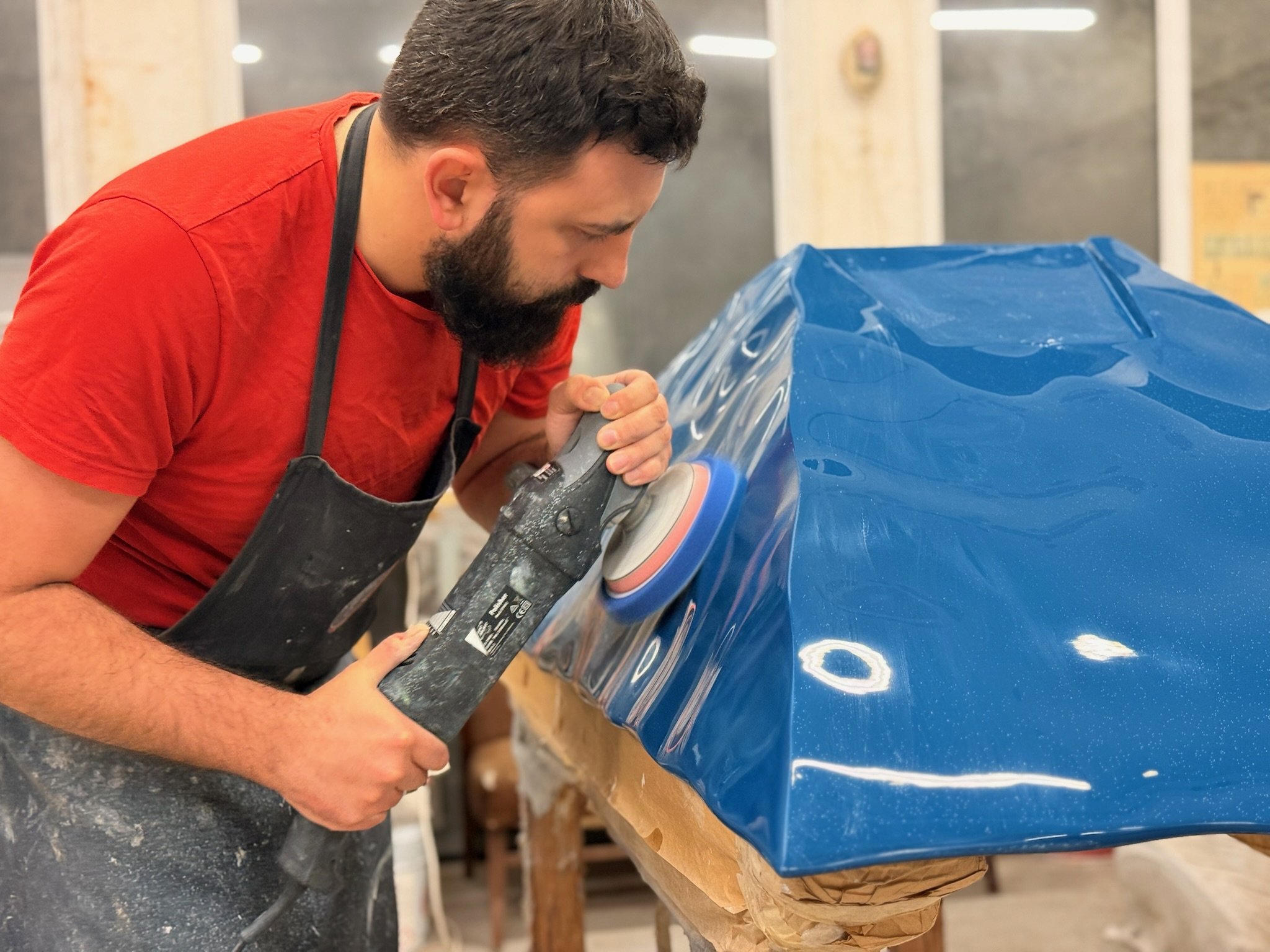 A man is polishing a blue car piece of art a power buffer in a workshop or garage.
