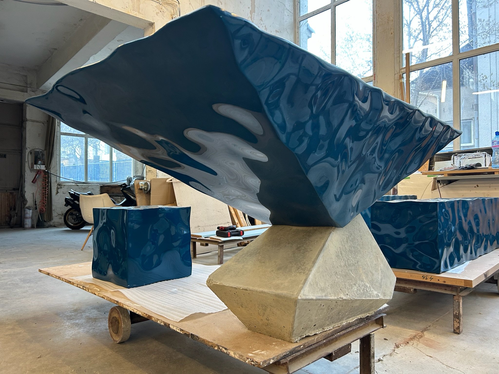 Large blue abstract sculpture with a glossy finish, supported by a concrete pyramid base, placed on a wooden platform inside an artist's studio.