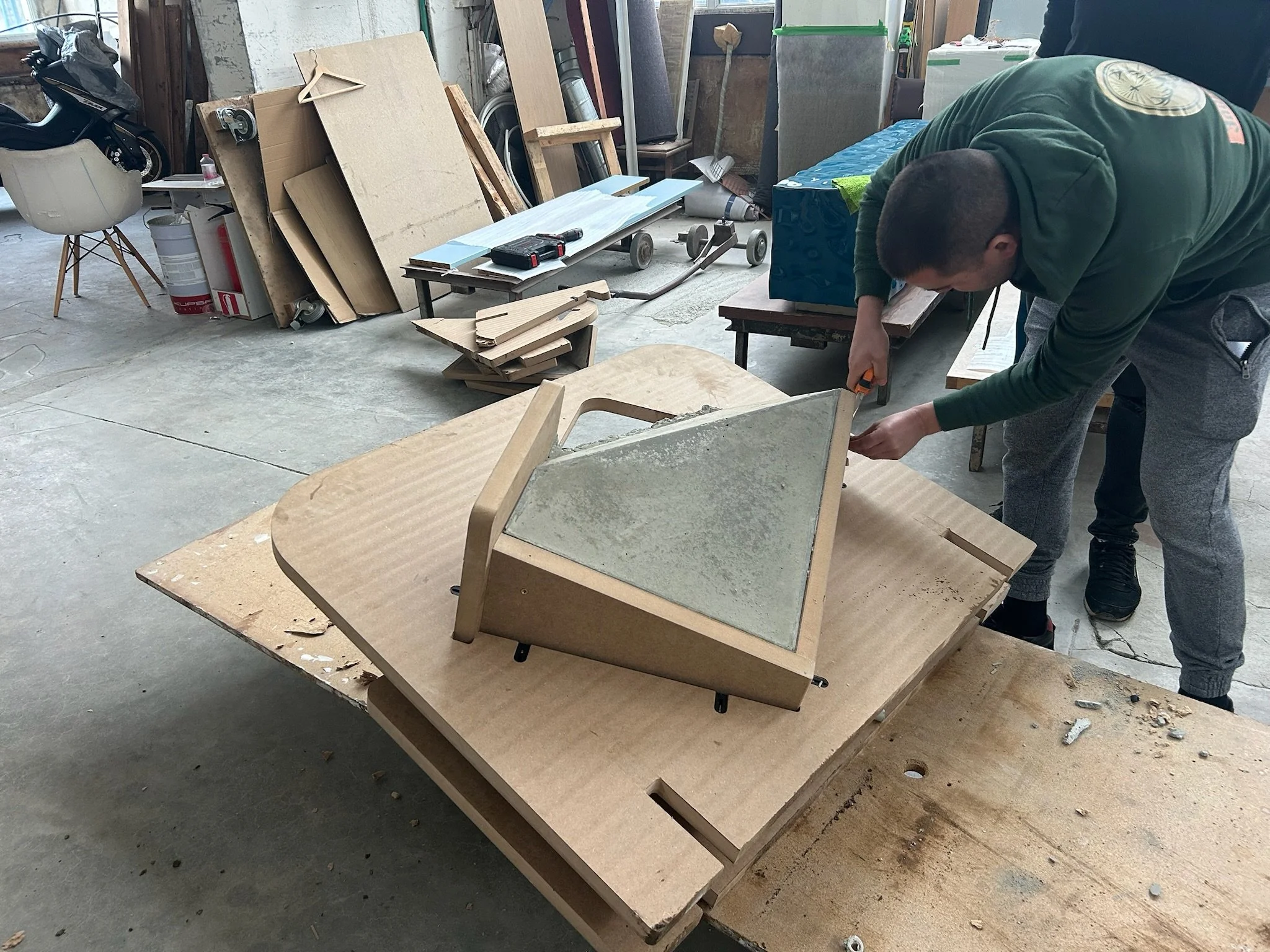 A person assembling a wooden and glass display case in a workshop, surrounded by woodworking supplies and tools, with various wood pieces and equipment in the background.